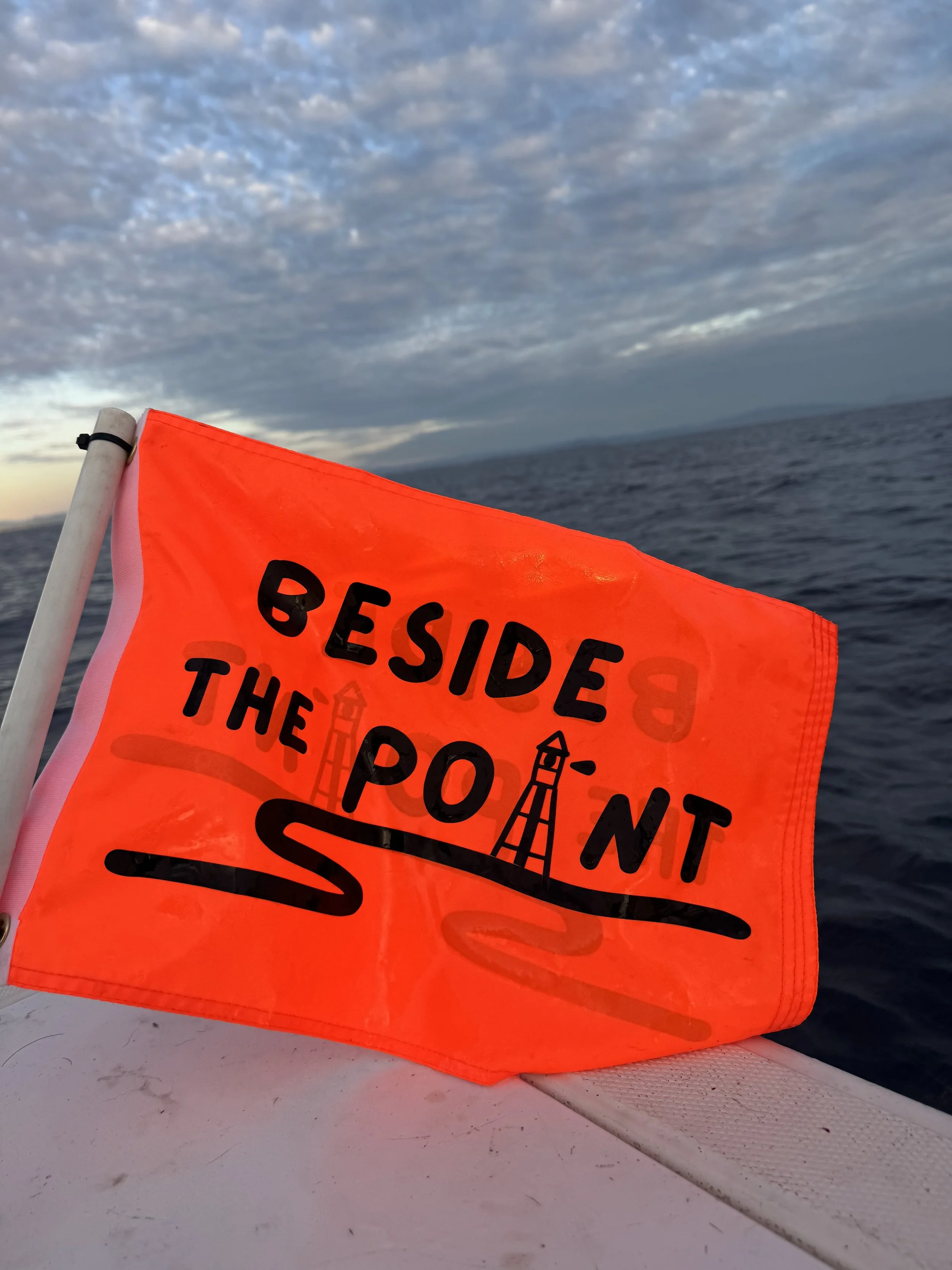 An orange safety flag with black text and lighthouse graphics, reading 'Beside the Point,' attached to a boat with ocean and cloudy sky in the background.