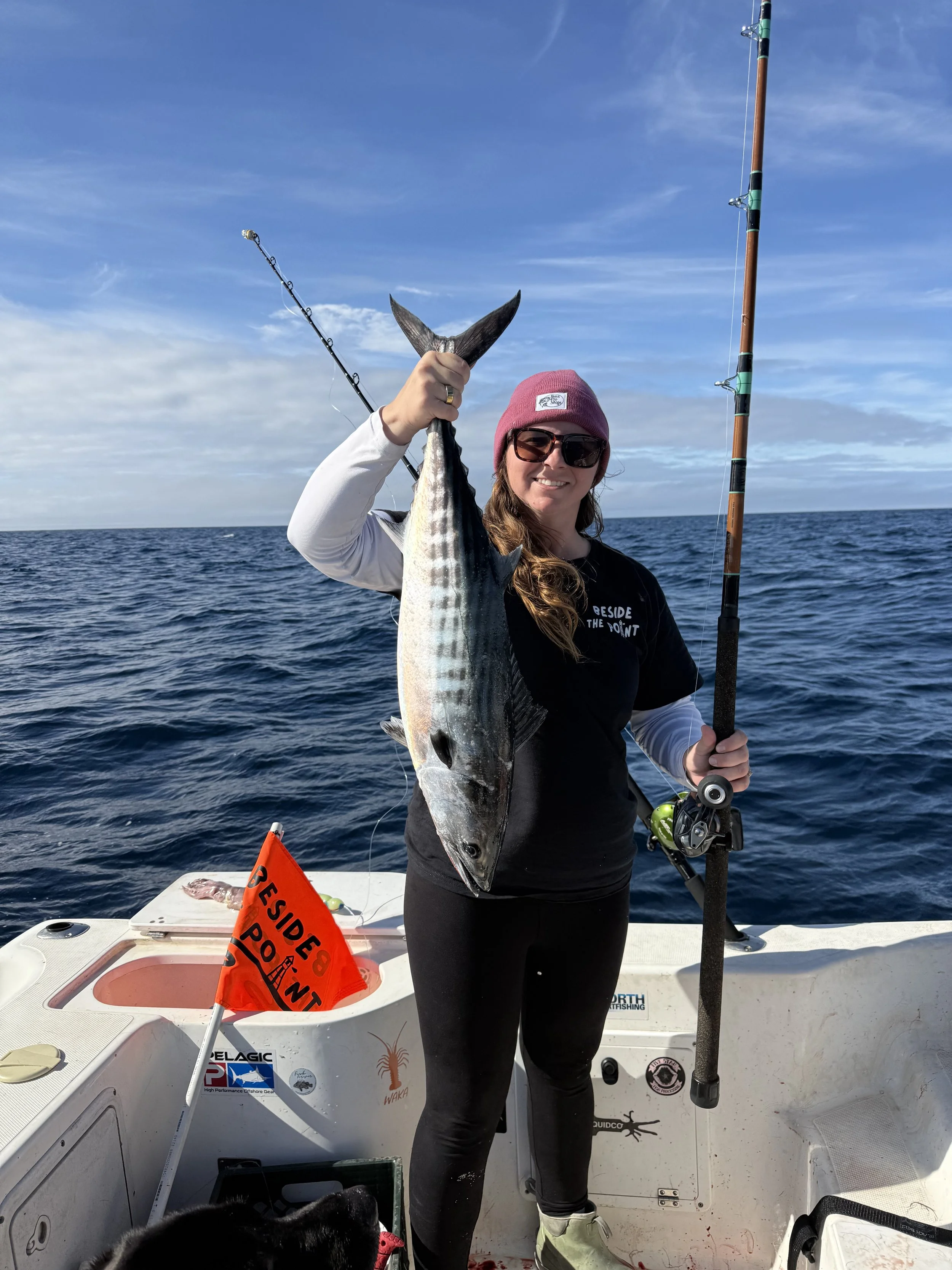 A woman holding a large fish on a boat in the ocean, with fishing rods and a small red flag that reads 'Beside the Point' in the background.