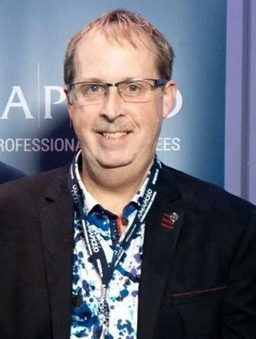 Middle-aged man with glasses, wearing a dark blazer and a patterned shirt, standing in front of a blue background with partial text.