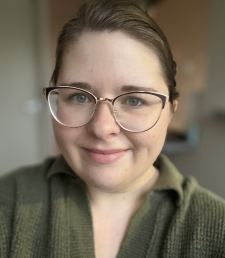 A young woman with glasses, smiling, wearing a green sweater, and having brown hair pulled back.