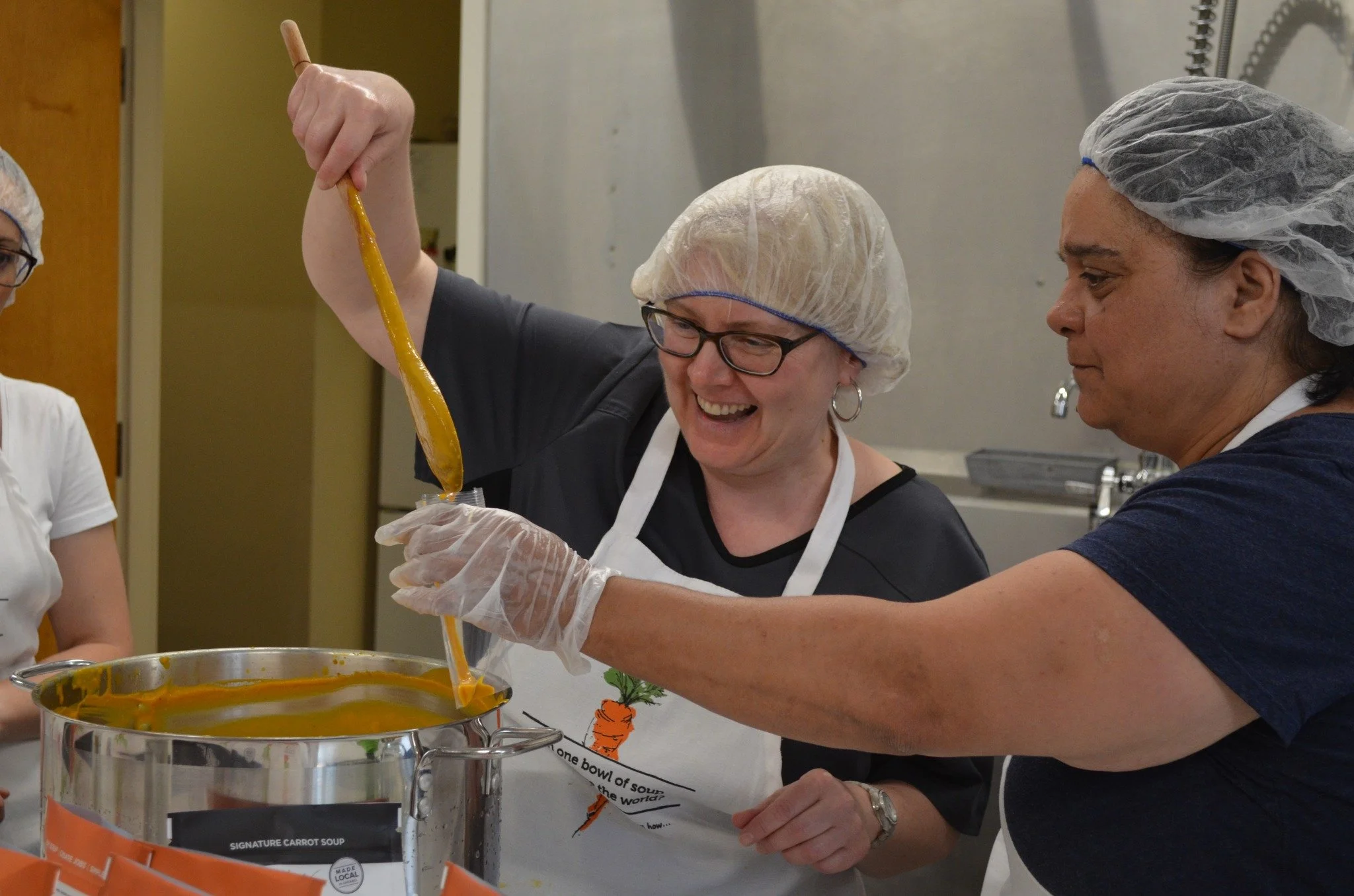 Two women wearing hairnets and aprons preparing soup in a commercial kitchen.
