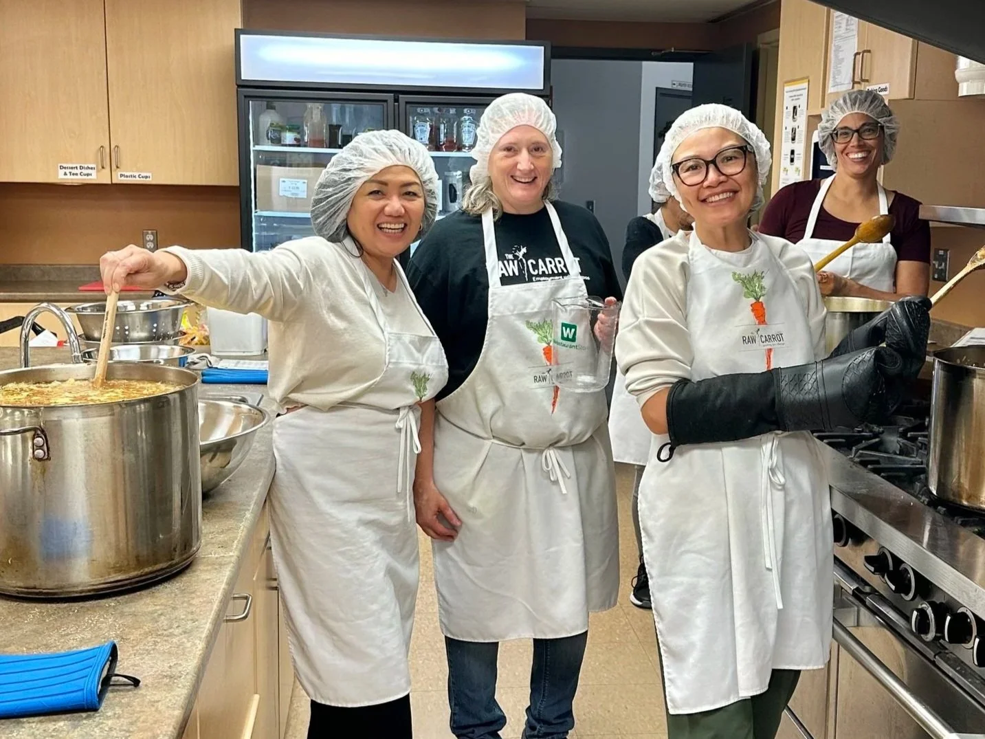 Five women in hairnets and aprons cooking in a kitchen, smiling at the camera. One woman is stirring a large pot of soup or stew; others are holding cooking utensils or standing nearby.