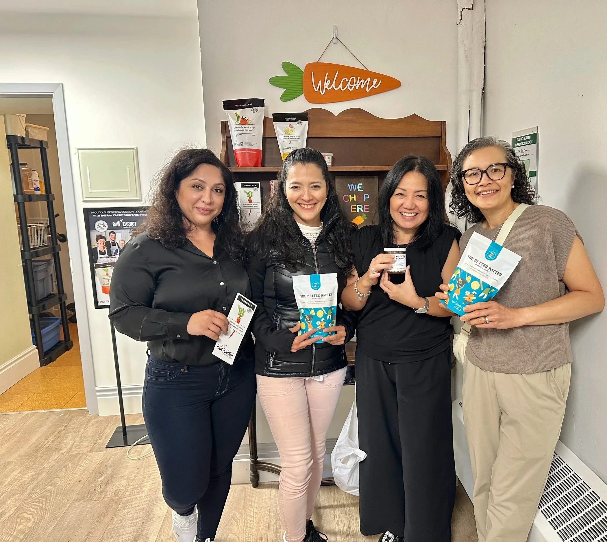 Four women standing indoors, smiling, holding food products and small jars, in front of a wooden shelf with food items and a