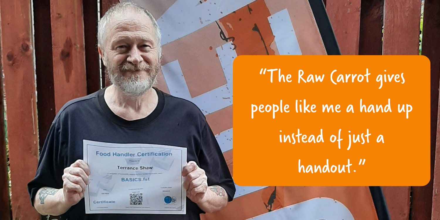 A man with a beard and tattoos holding a food handler certification certificate, standing outdoors in front of a wooden fence with an orange and gray banner. He is smiling and wearing a dark t-shirt.