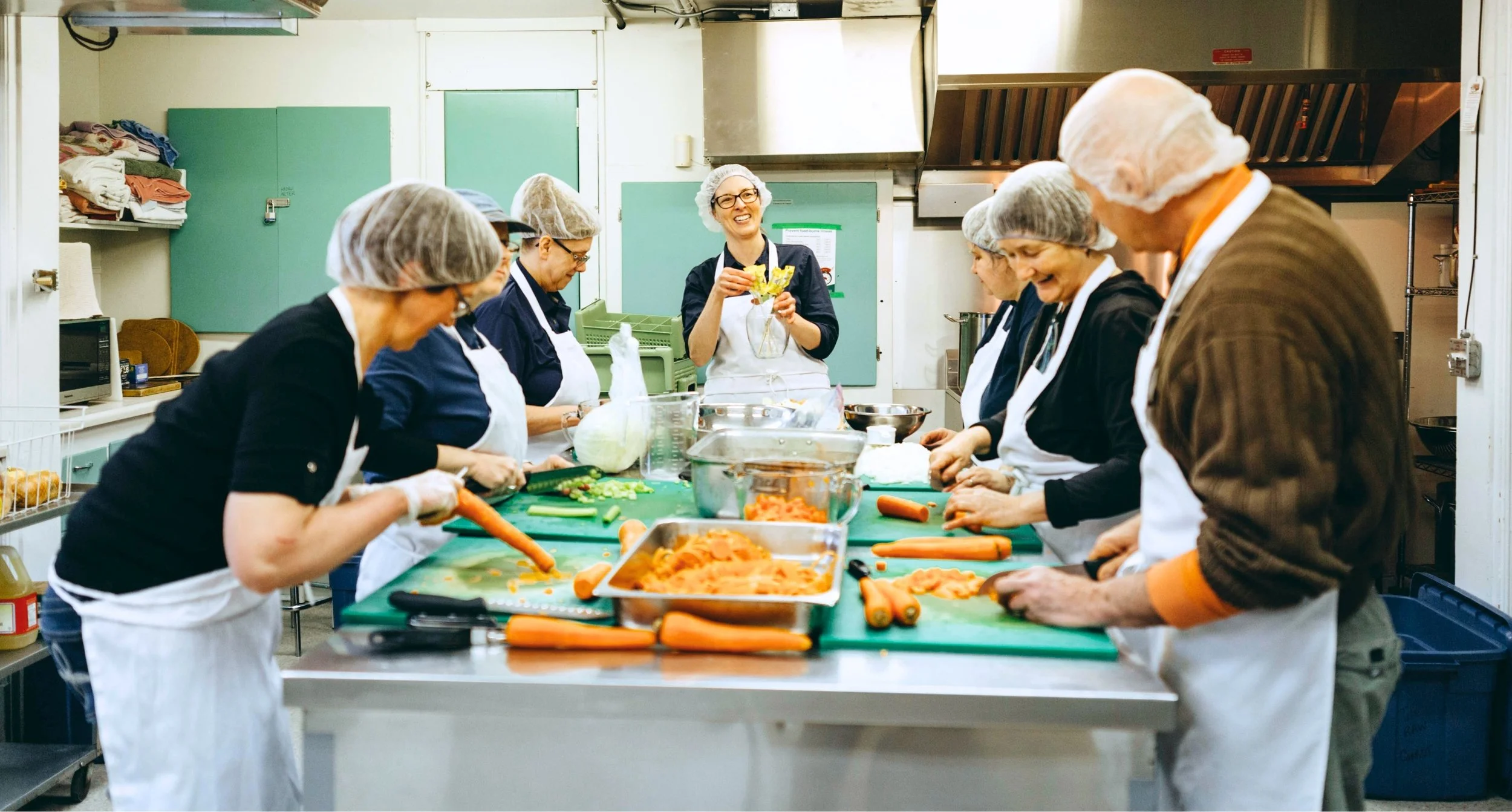 A group of people wearing hairnets and aprons preparing and chopping vegetables in a commercial kitchen, with one woman smiling and holding yellow vegetables while others focus on their tasks.