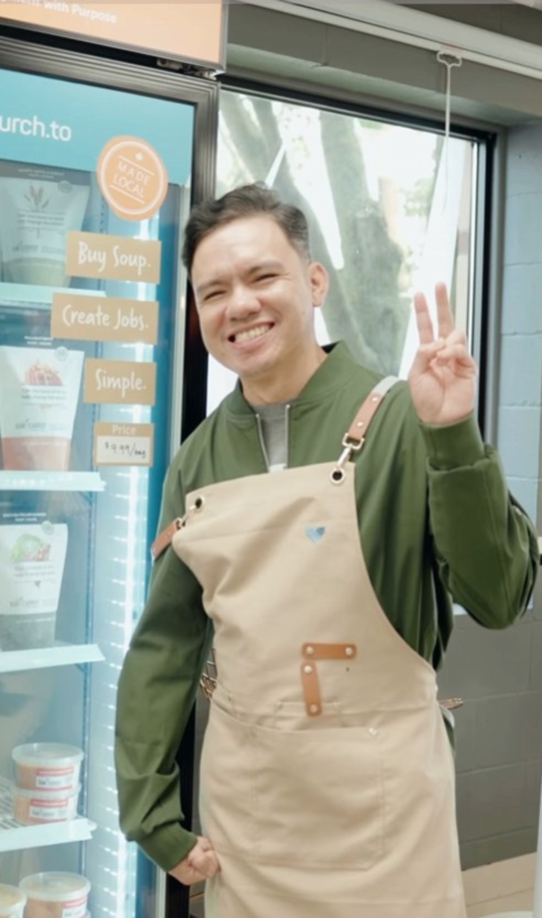 A young man wearing a green jacket and beige apron with a small triangular logo, standing in front of a refrigerated display, smiling and making a peace sign with his right hand.