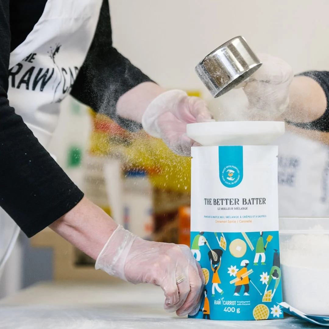 Person wearing gloves pouring flour from a metal sifter into a large bag of flour labeled 'The Better Batter' with colorful illustrations on a kitchen countertop.