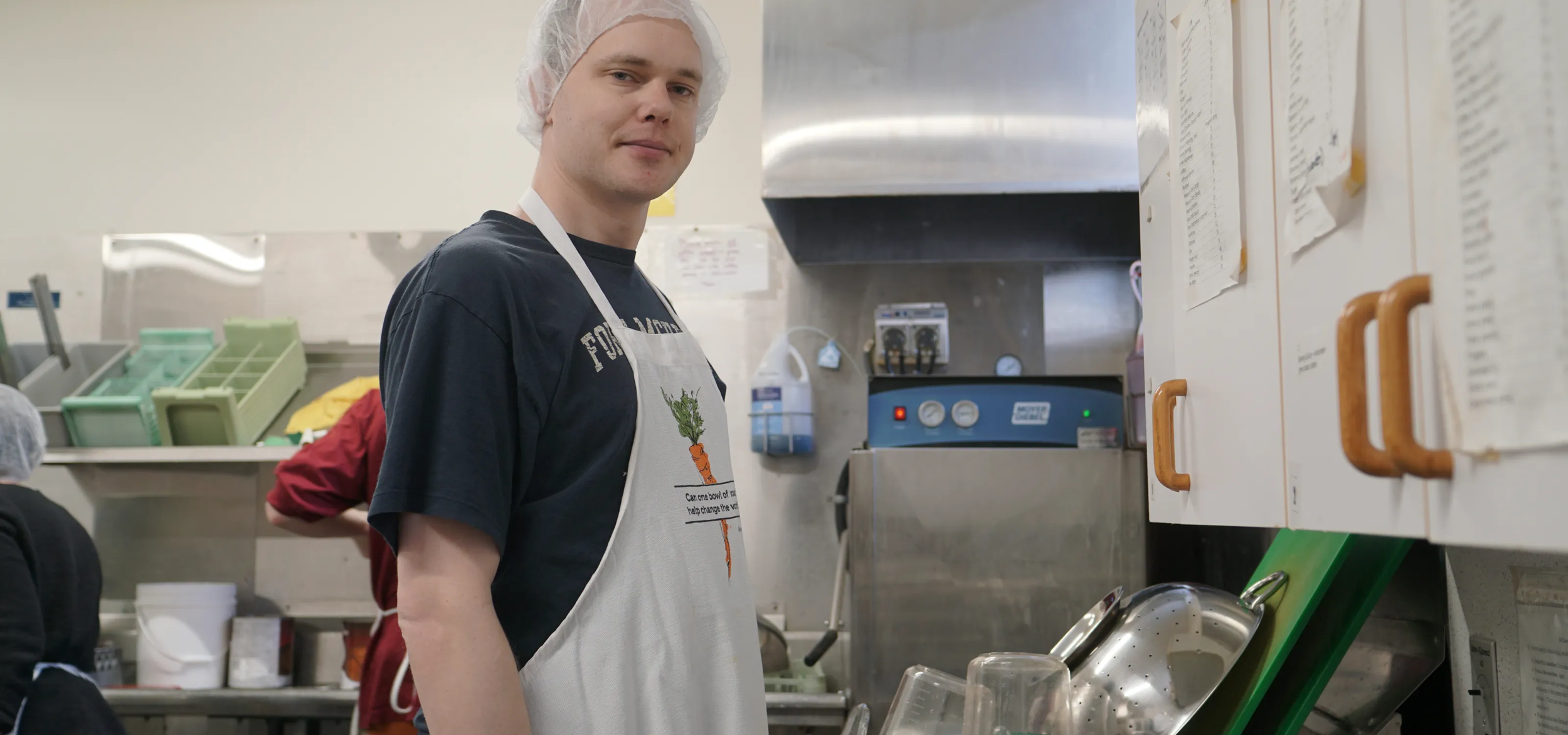 A young man wearing a hairnet and apron with a carrot design stands in a kitchen, looking at the camera.