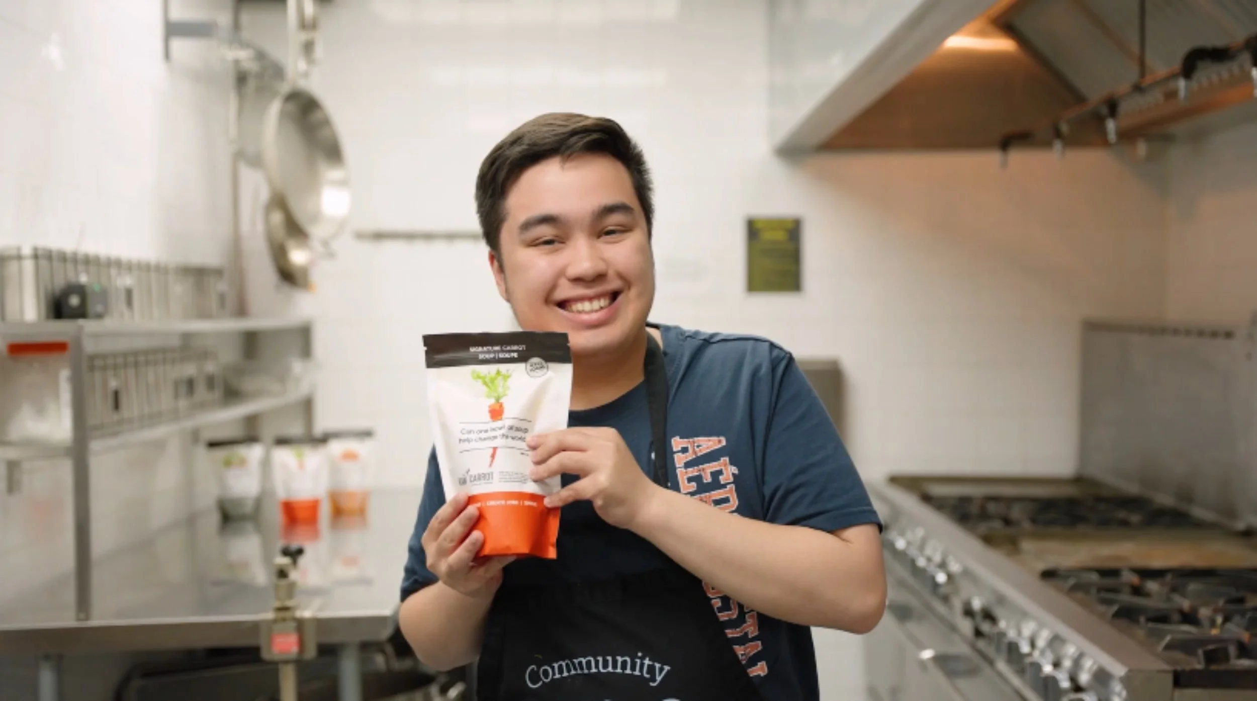 A smiling young man wearing a dark apron in a commercial kitchen holding a package of carrot soup mix.