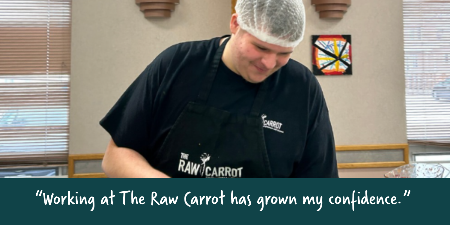 A person wearing a hairnet and black apron with The Raw Carrot logo, working in an indoor setting with wooden blinds and artwork on the walls.