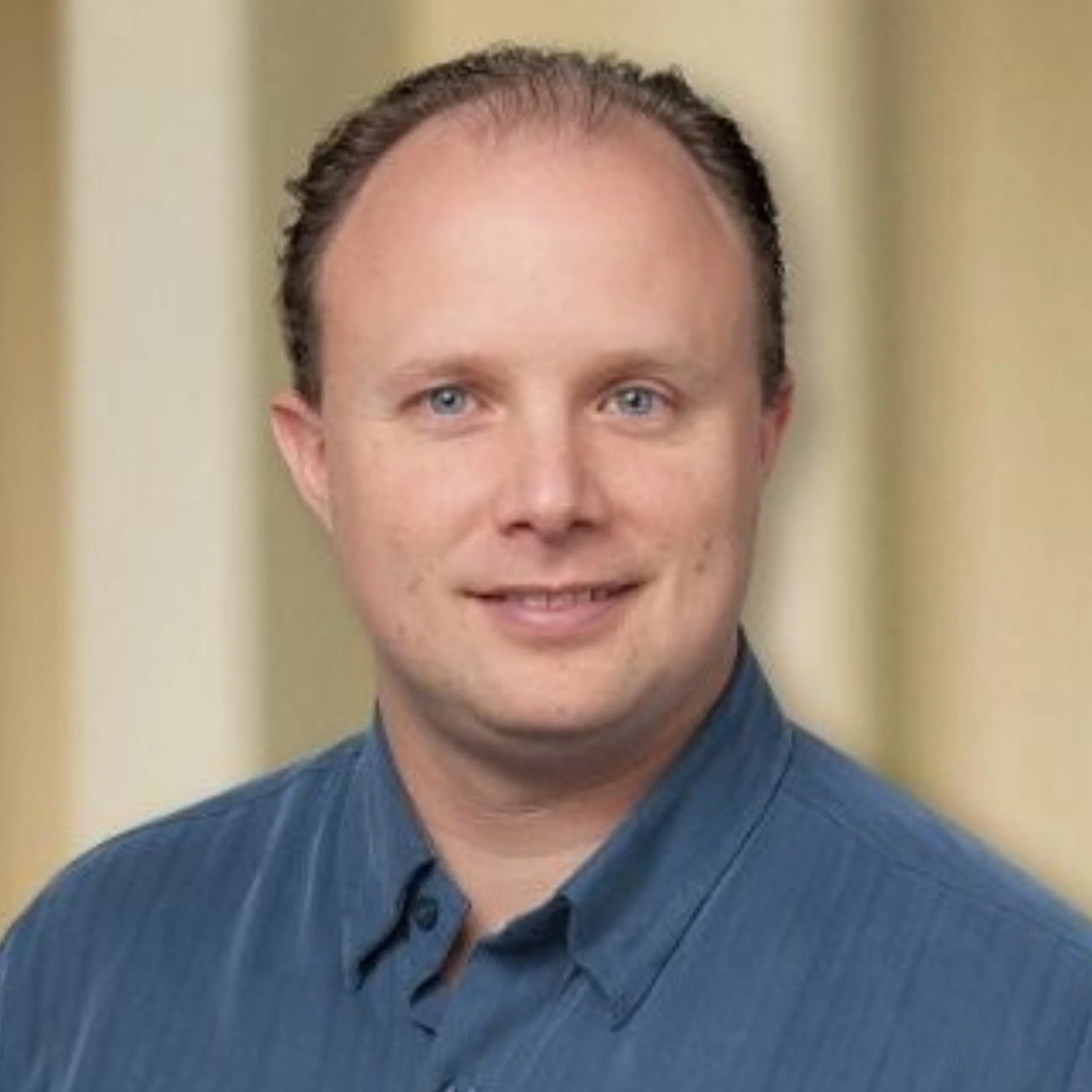 A smiling man with short dark hair and blue eyes, wearing a light blue collared shirt, posed against a blue wall.