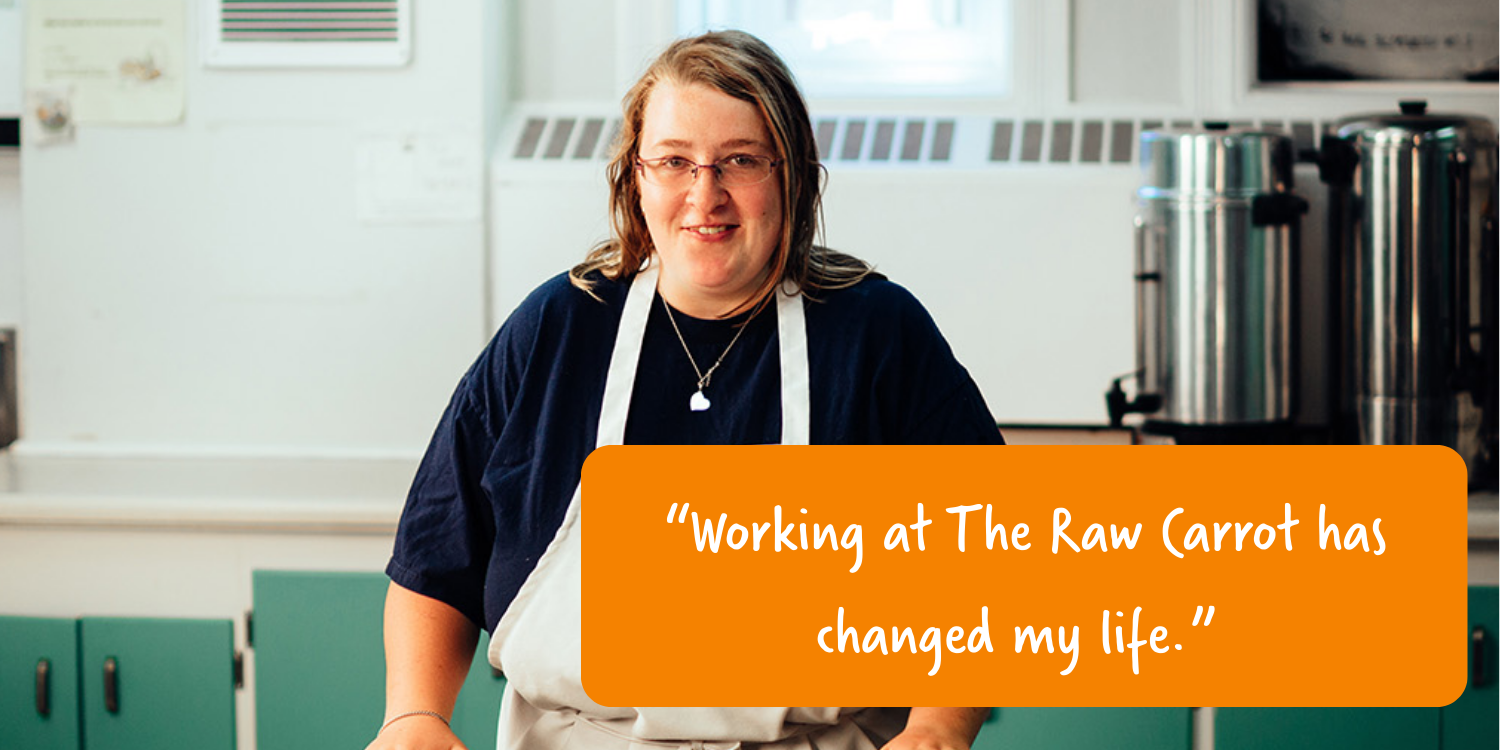 A woman wearing glasses, a black shirt, and a beige apron stands in a kitchen with teal cabinets, white walls, and stainless steel appliances. There is an orange text box with white handwritten style font that reads, 'Working at The Raw Carrot has changed my life.'