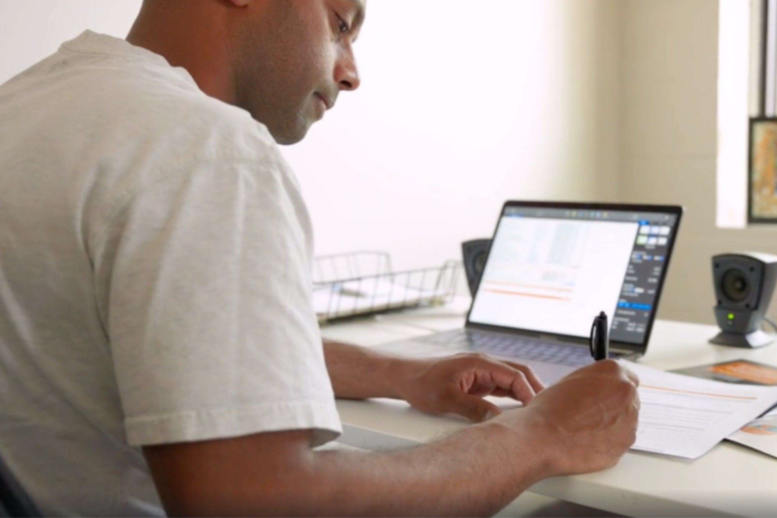 A man working at a desk with a laptop, a pen, and papers, two speakers are on the desk, and a window in the background.