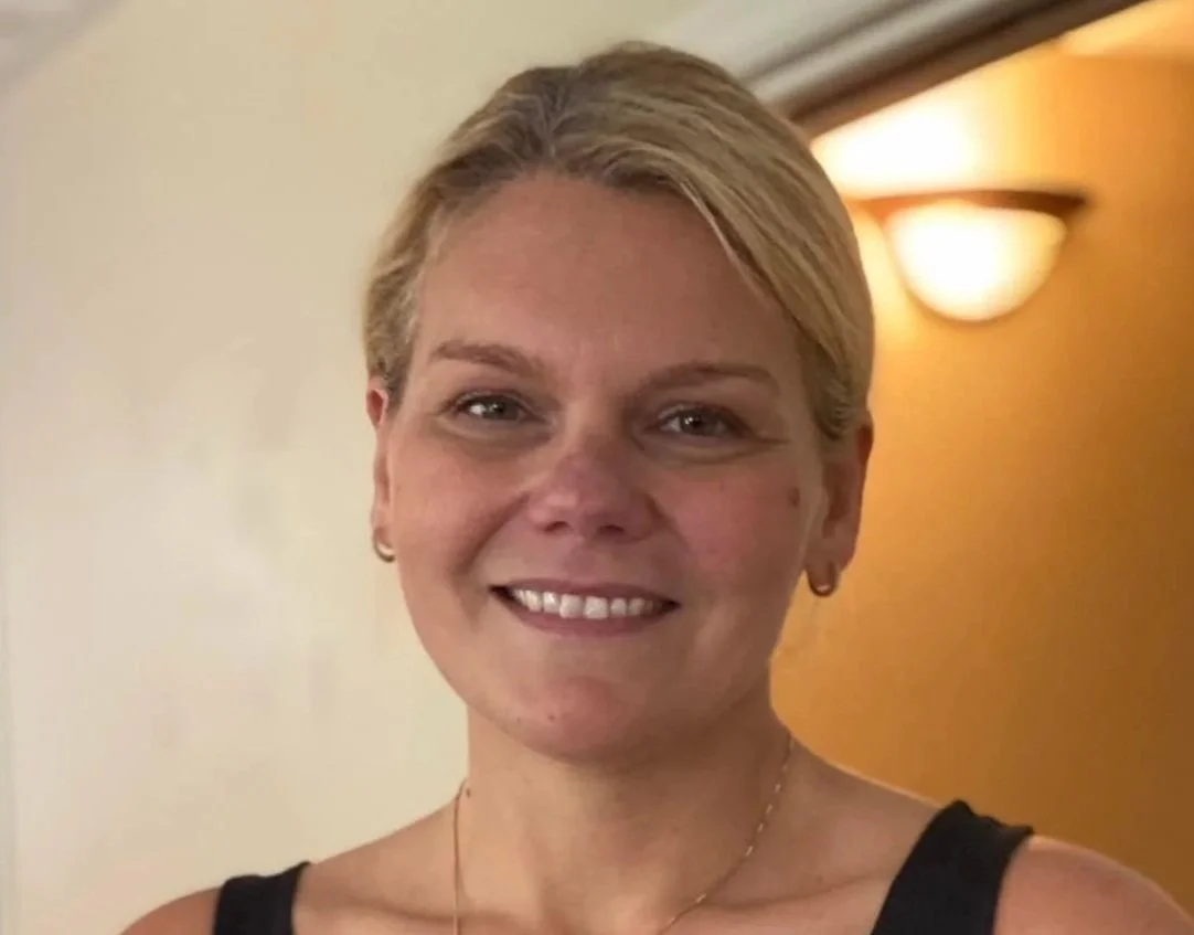 A smiling woman with short blonde hair, wearing a black top and small earrings, standing indoors in front of a cream wall with a yellow accent wall and a wall-mounted light fixture in the background.