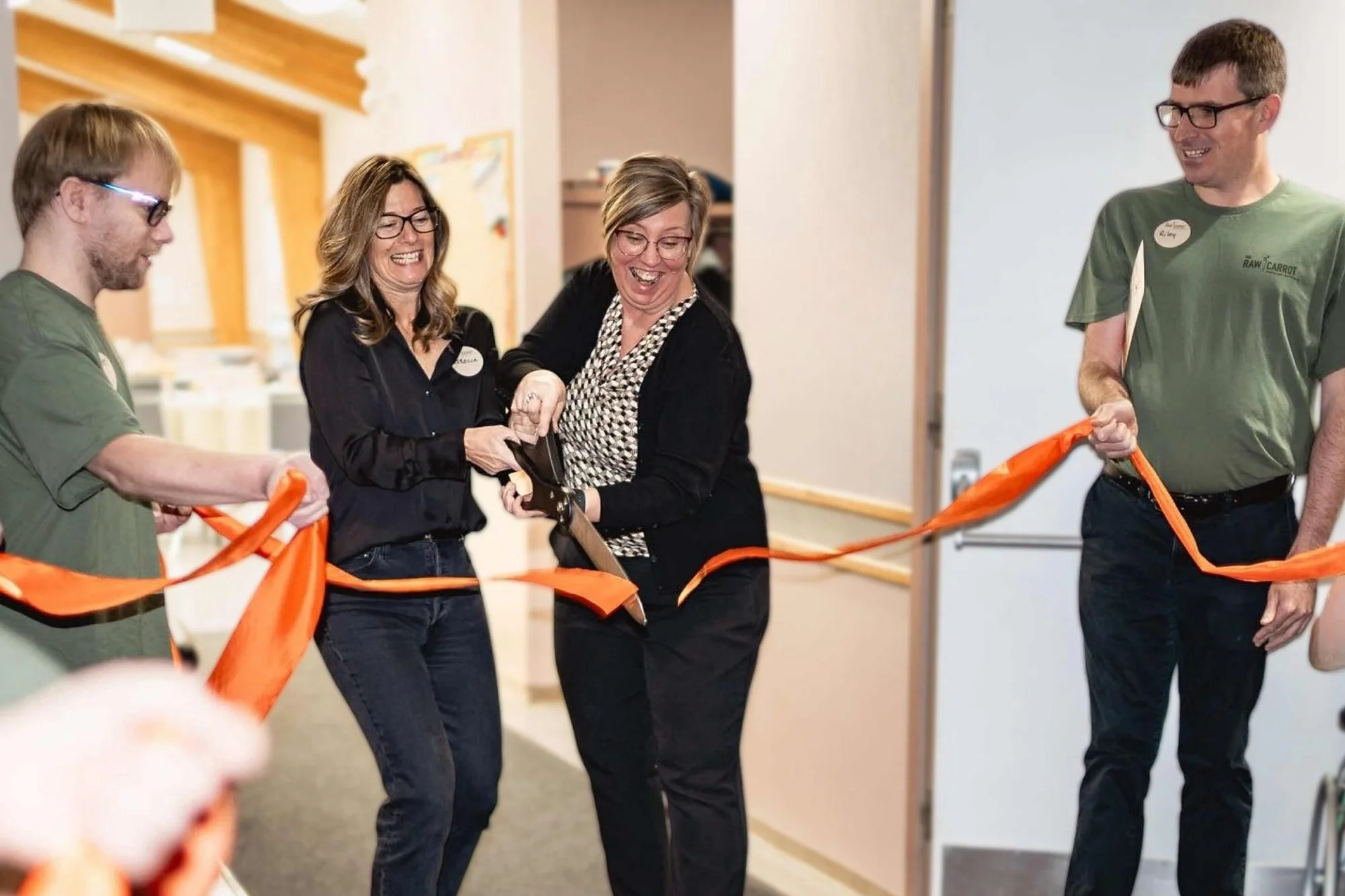 Four people, three adults and one young man, smiling and participating in a ribbon-cutting ceremony. The woman in the middle is using scissors and the others hold an orange ribbon.