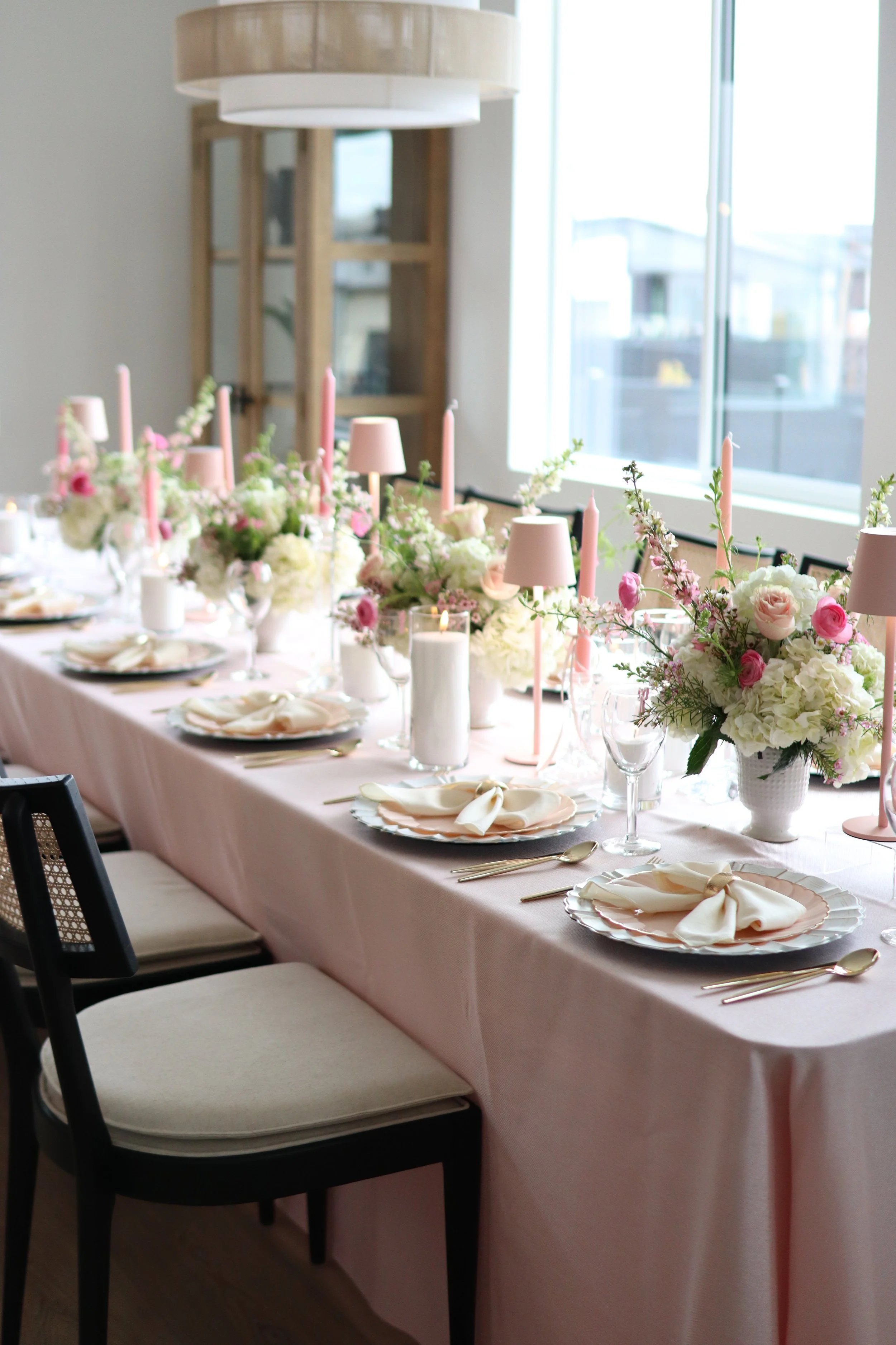 A long dining table decorated for a special event with pink and white floral arrangements, candles, pink candles, white napkins, and elegant dinnerware, set near large windows in a bright, modern room.