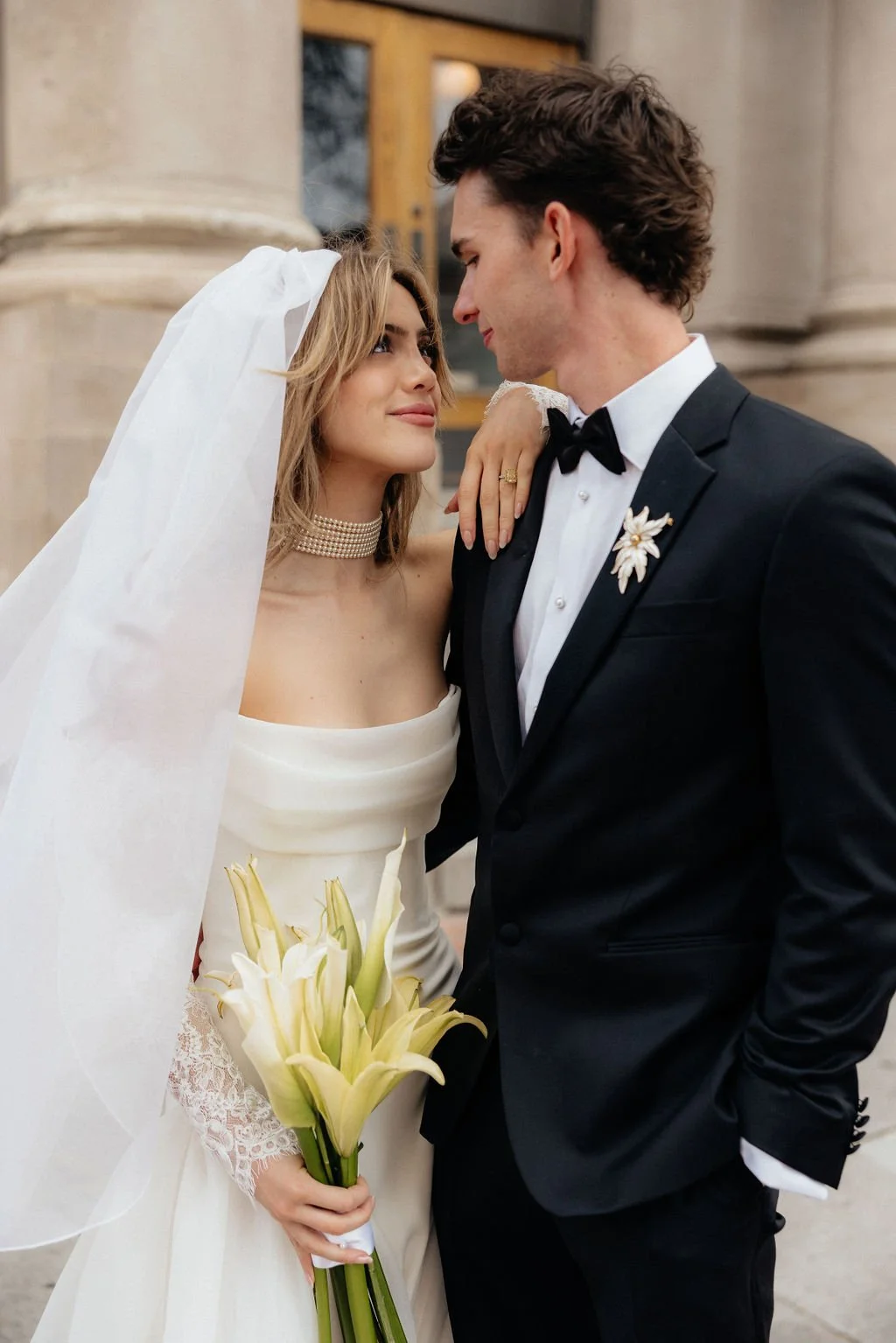 A bride in a lace gown holding a sophisticated bouquet of crisp white lilies, gazing at her groom against a backdrop of classical stone columns.