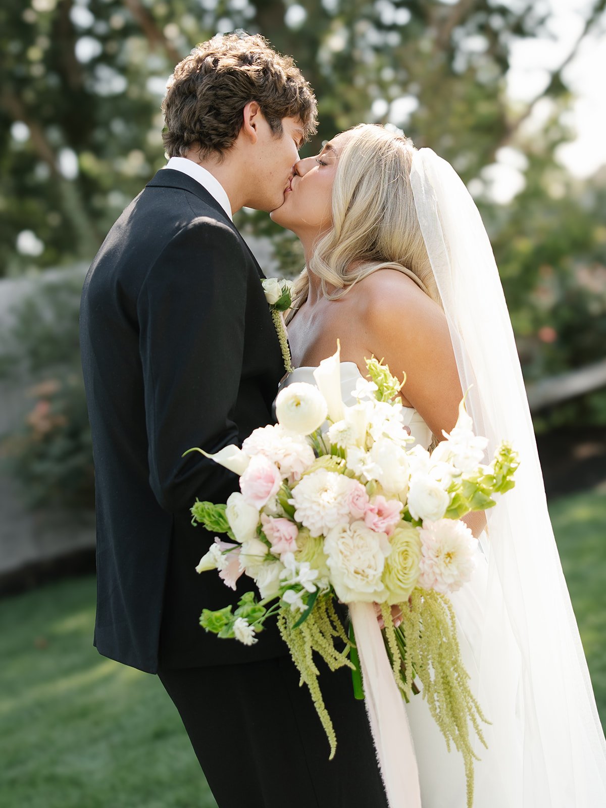A bride and groom kissing outdoors on their wedding day, with the bride holding a bouquet of white and pink flowers.