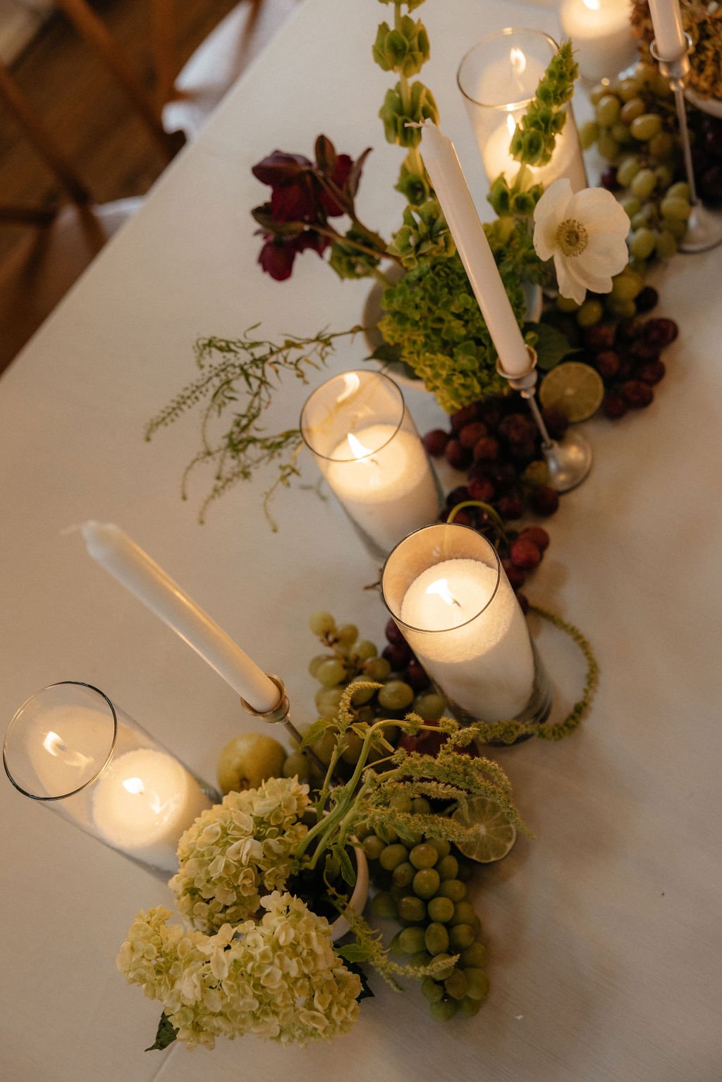 A luxury wedding tablescape featuring an artistic floral runner with deep burgundy blooms and verdant green hydrangeas, accented by glowing pillar and taper candles in glass hurricanes.