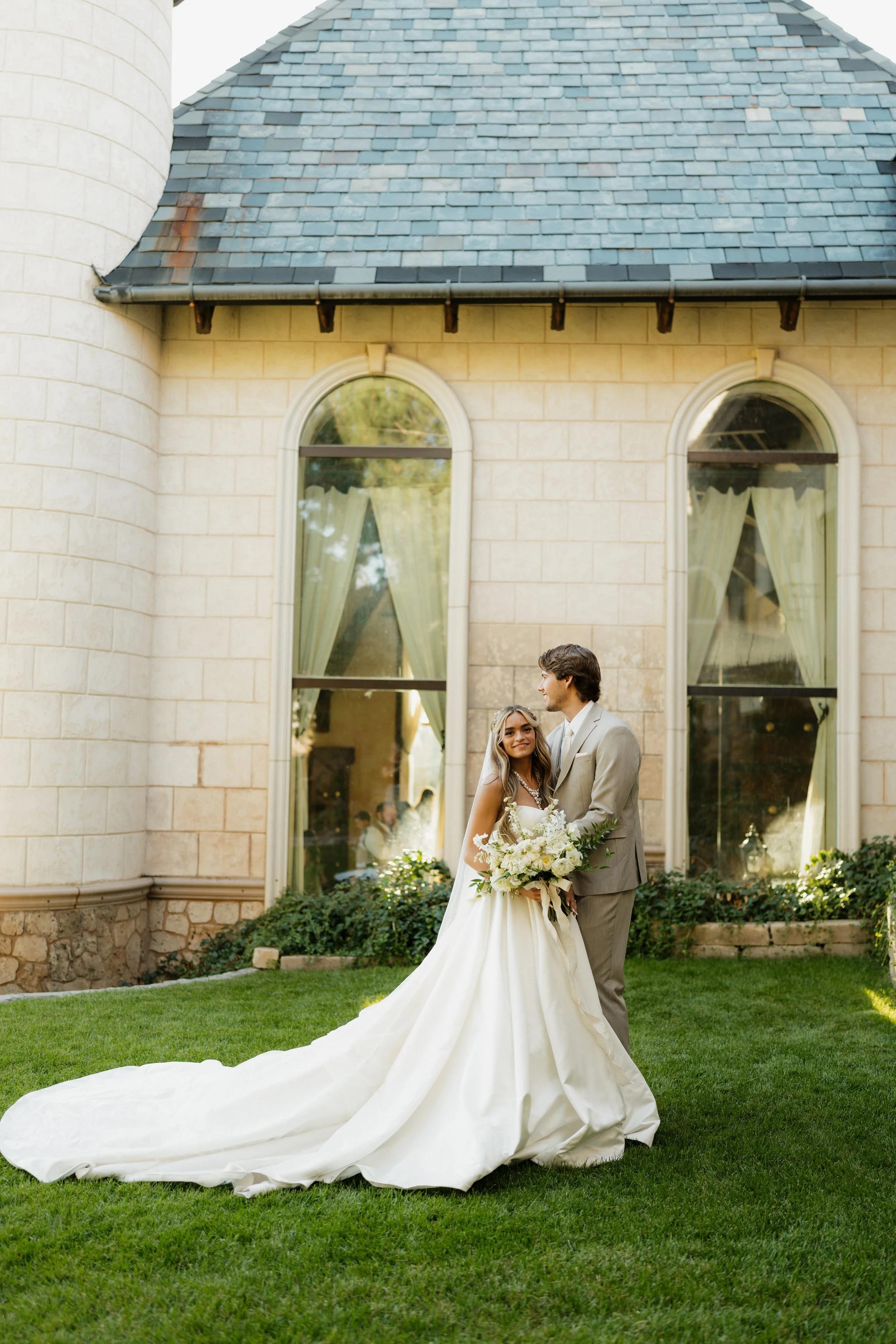A bride and groom standing outdoors in front of a stone building with large arched windows. The bride is wearing a white wedding gown with a long train, holding a bouquet, and the groom is wearing a light-colored suit. They are smiling at each other,