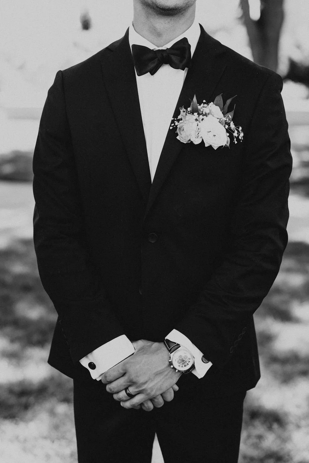 A man in a tuxedo with a bow tie and boutonniere, standing outdoors with his hands clasped, wearing a wristwatch and wedding band.