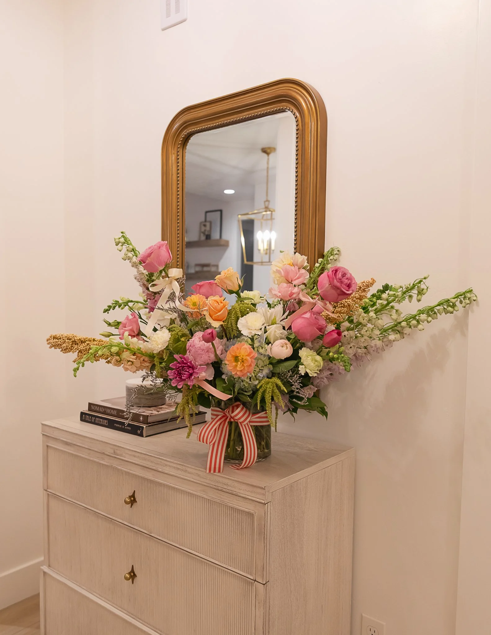 A bouquet of pink, peach, and white flowers in a glass vase with a pink and white striped bow, placed on a white dresser in front of a large gold-framed mirror.