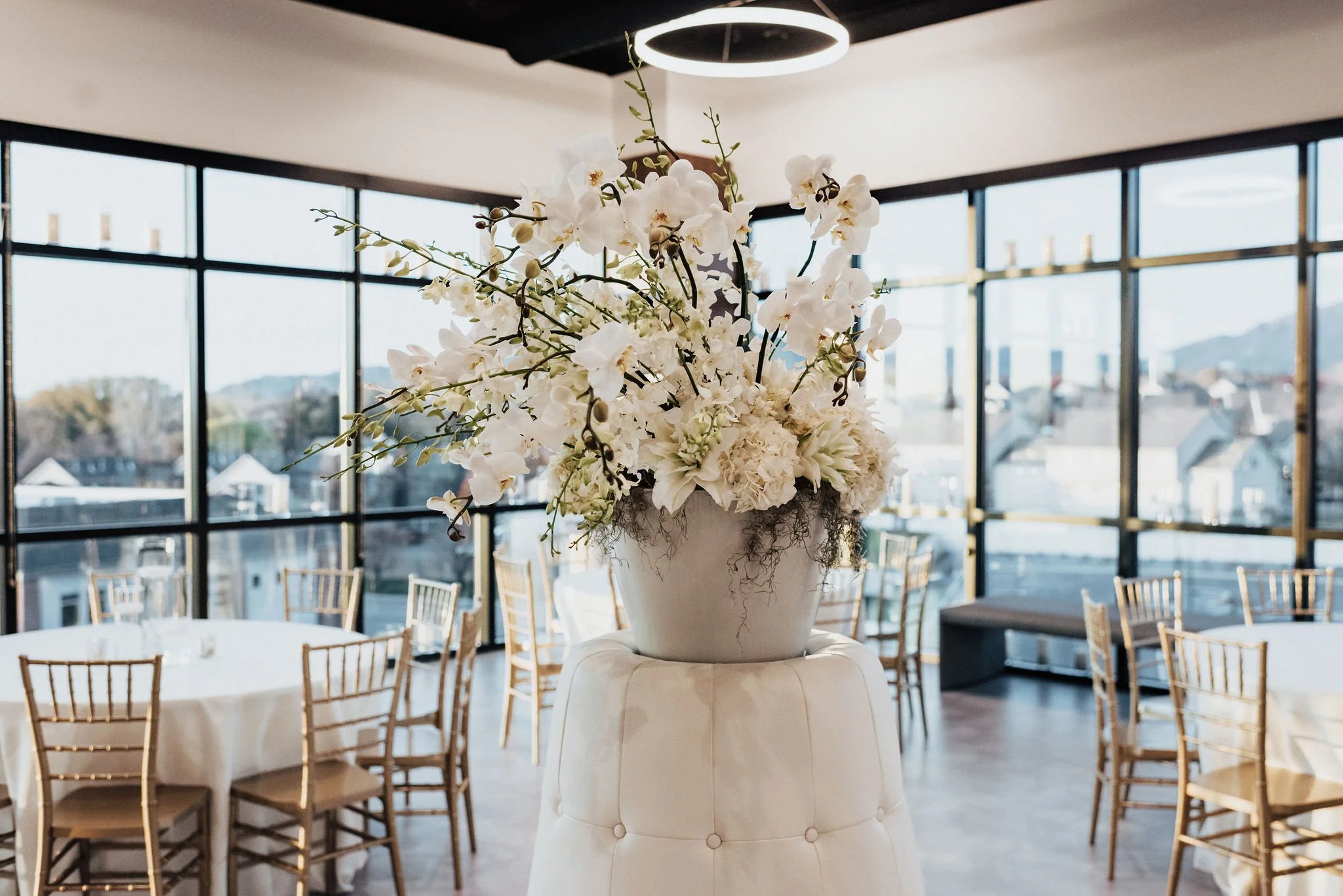 Elegant flower arrangement with white orchids and other white flowers in a gray vase on a white ottoman, inside a bright, modern event space with large windows and gold chairs.