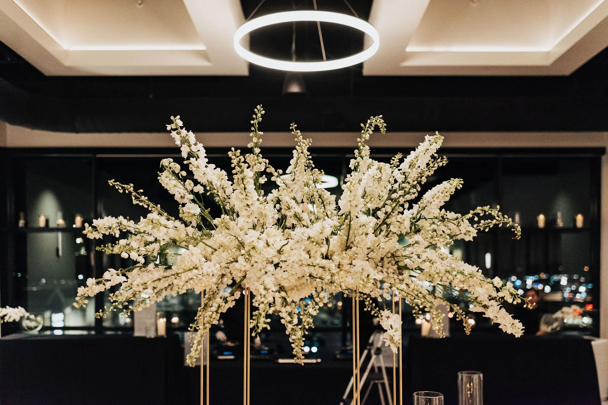 A large white floral arrangement on a tall stand in a modern, dimly-lit room with dark walls and ceiling, with city lights visible through the windows at night.