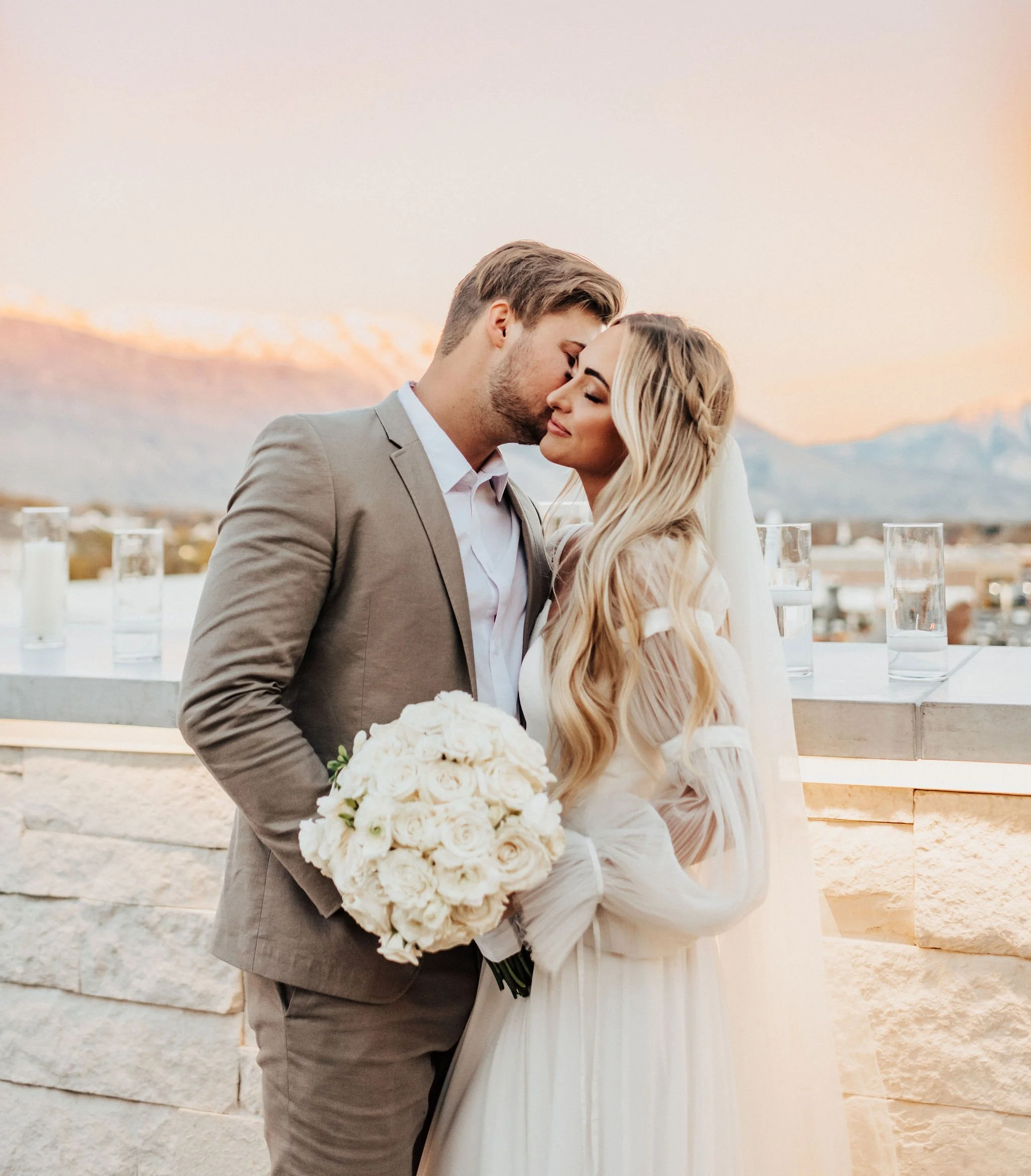 A couple embracing during a wedding ceremony at sunset, with the man in a gray suit holding a bouquet of white roses, and the woman in a white dress with long flowing hair, against a backdrop of mountains and a soft pastel sky.