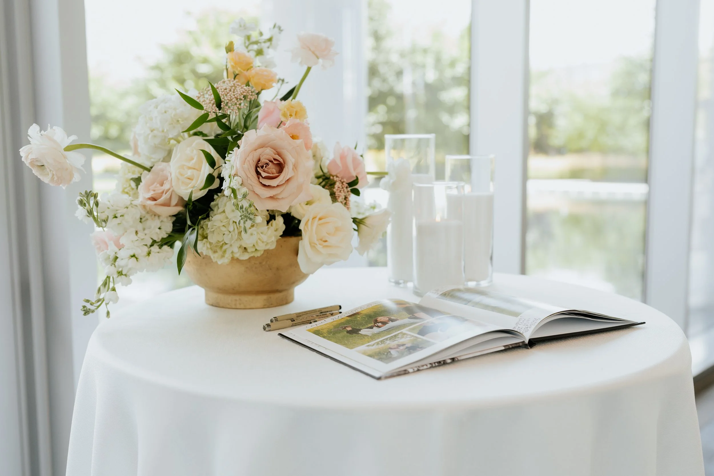A round table with a white tablecloth holds a large bouquet of pastel pink and white flowers, an open photo album, three black and silver pens, and three tall glass candles, with a bright window and greenery outside in the background.