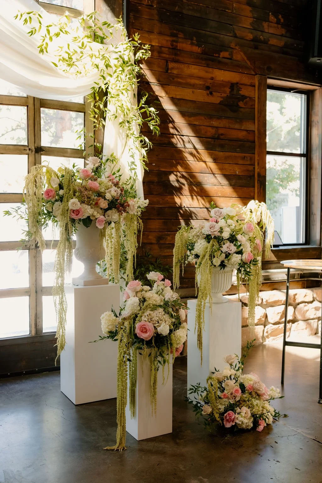 Elegant floral arrangement with white and pink roses, white hydrangeas, and cascading greenery in white vases, set against rustic wooden walls and large windows with natural light.