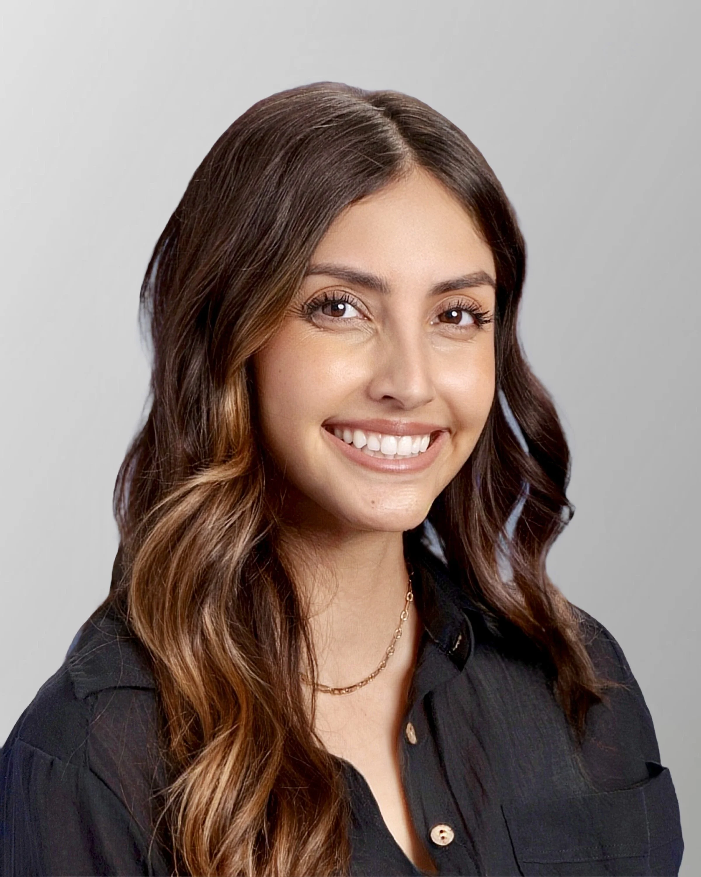 Portrait of a young woman with long wavy brown hair, smiling, wearing a black blouse and gold necklace, against a gray background.