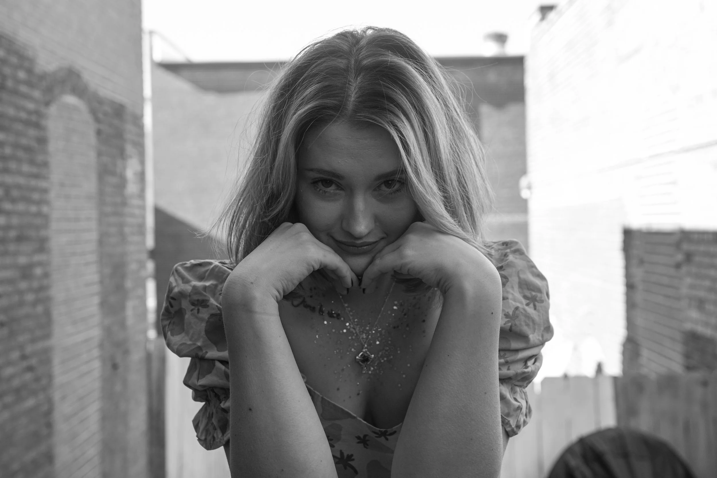 A woman with shoulder-length hair resting her chin on her hands, smiling slightly at the camera, outdoors on a sunny day with brick walls in the background.