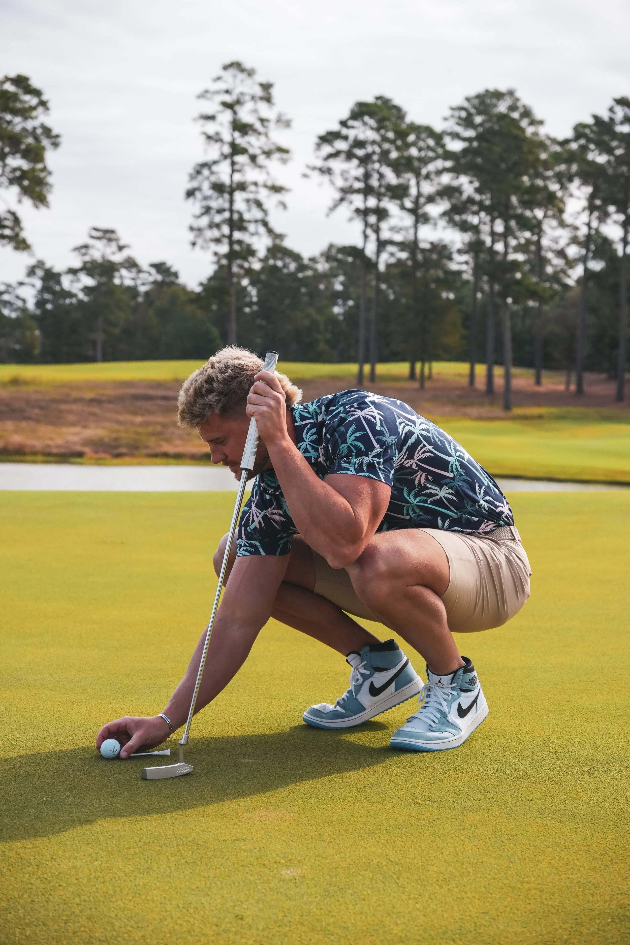 A man crouching on a golf green, placing a golf ball. He is holding a golf putter and is dressed in a patterned shirt, khaki shorts, and Nike high-top sneakers.