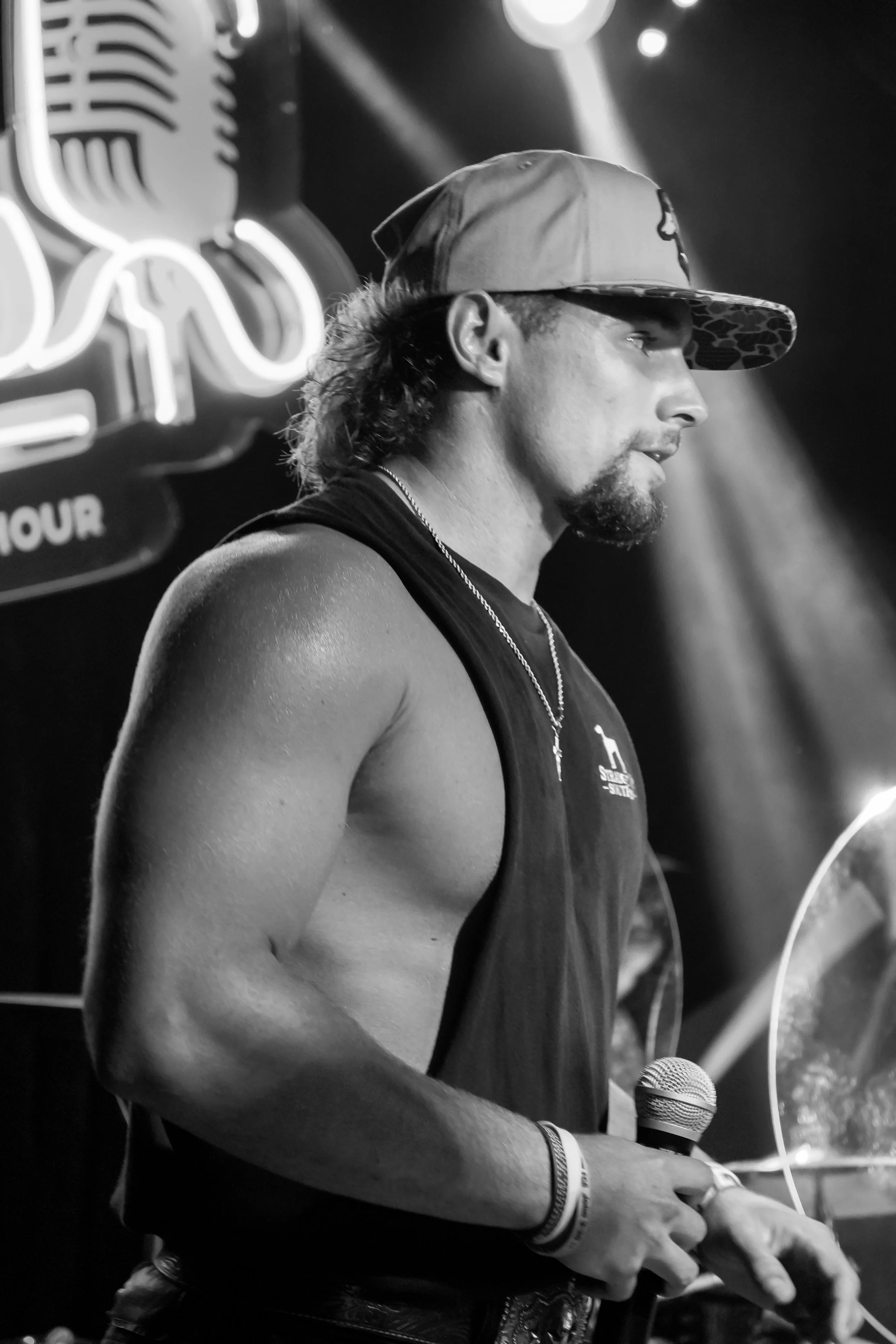 A man with a beard and mustache, wearing a cap and tank top, holding a microphone, on stage at a music venue, black and white photo.