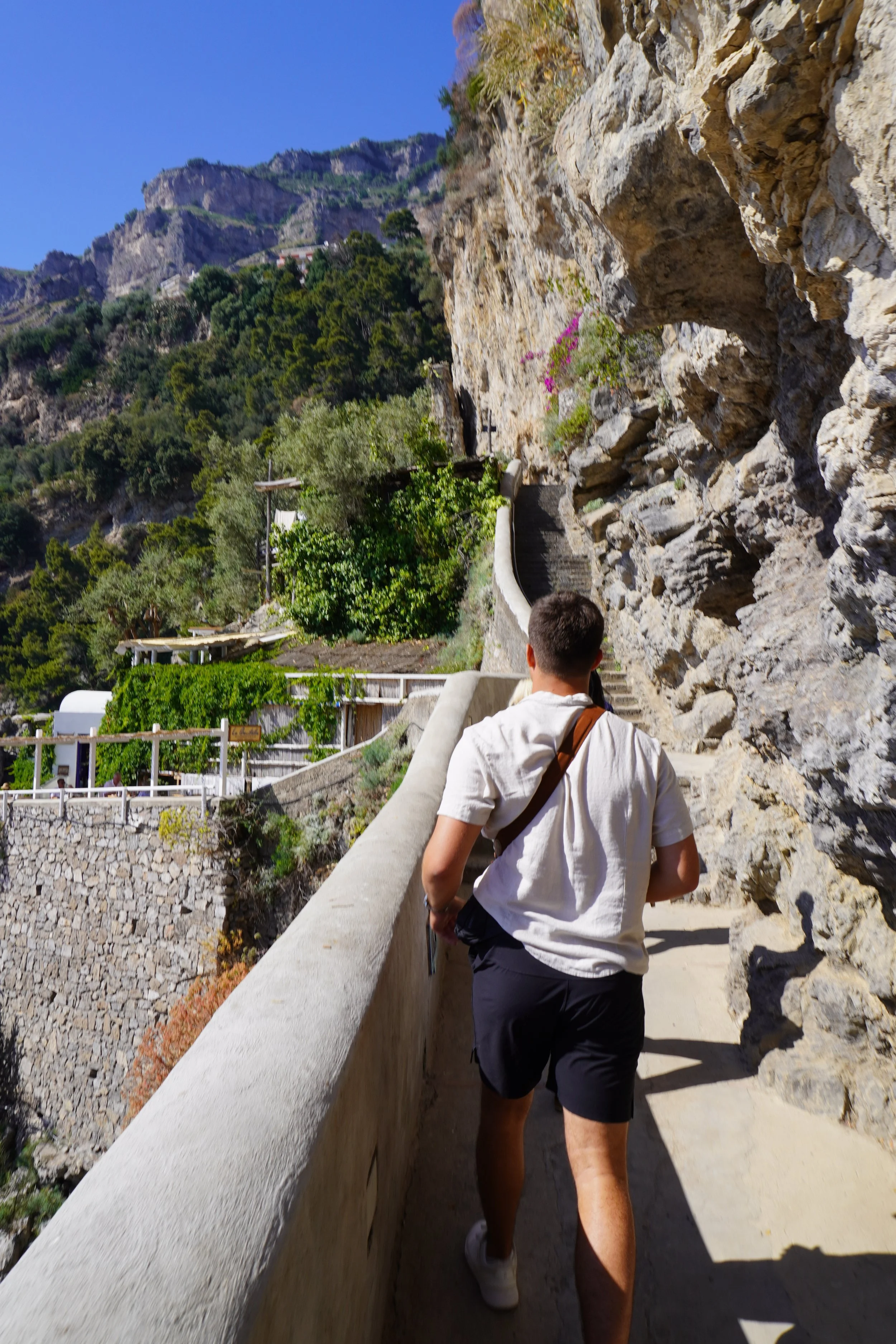 A person walking along a rocky mountain trail with a concrete railing on a sunny day.