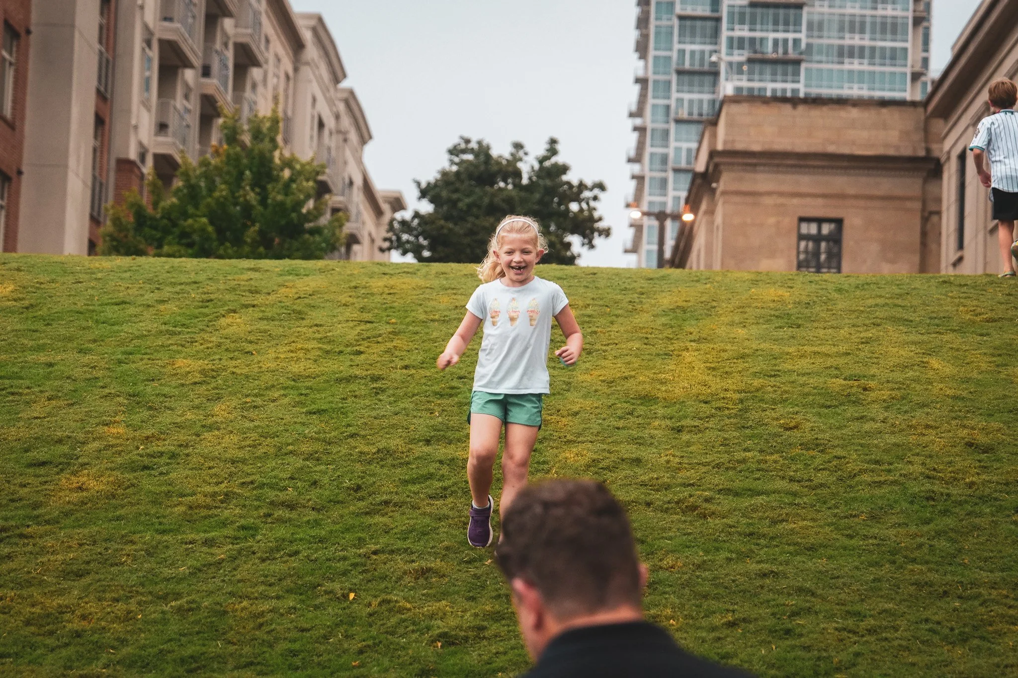 A young girl with a ponytail is running and smiling down a grassy hill in an urban park, with buildings and another child on the top of the hill in the background.