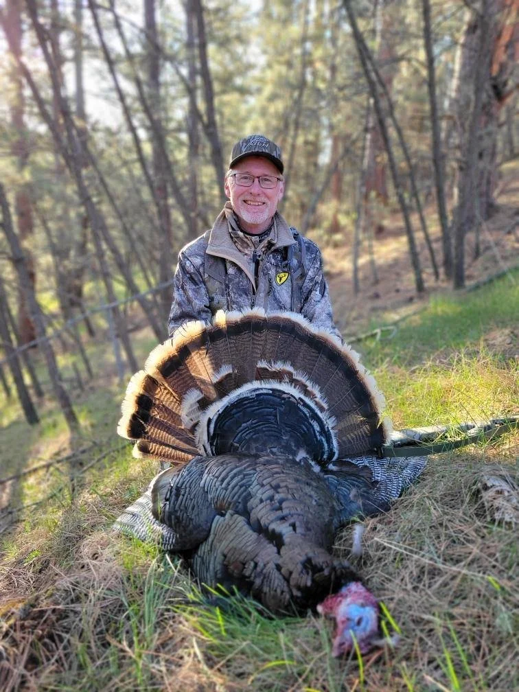 A smiling man in camouflage outdoor clothing and a cap kneels next to a large dead turkey on the ground in a forested area with trees and grass in the background.