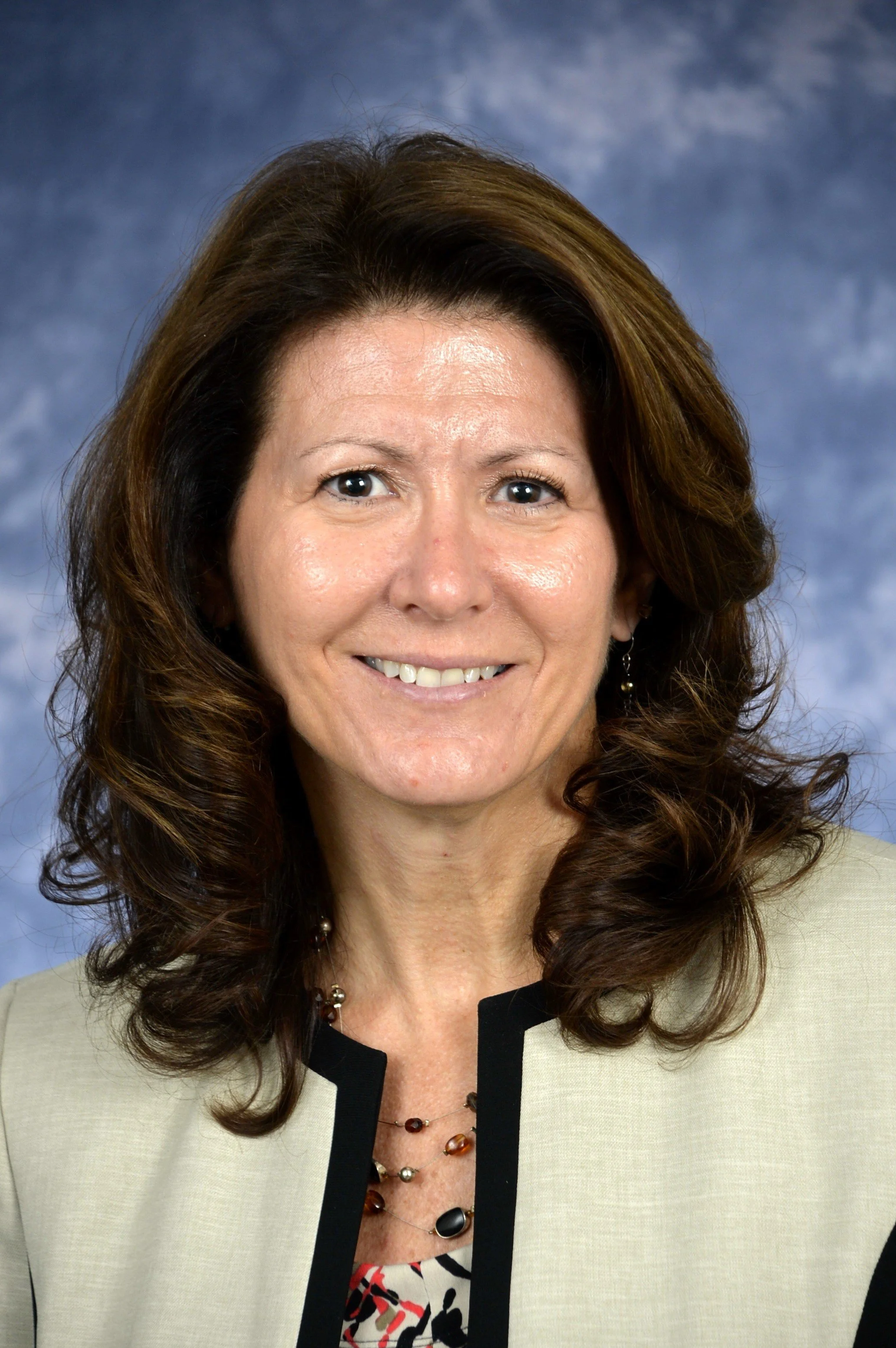A professional woman with brown, wavy hair wearing a beige blazer with black trim, a multicolored patterned top, a necklace, and earrings, smiling against a blue backdrop.