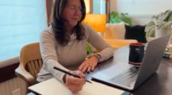 Woman sitting at a desk with a laptop, notebook, and pen, working in a bright room with a window and houseplants.