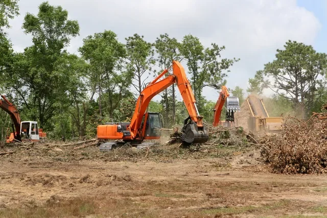 Construction site with orange excavators digging and moving dirt amid trees.