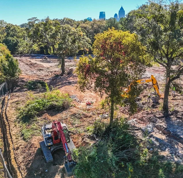 Construction site with excavators working among trees, with a city skyline visible in the background.