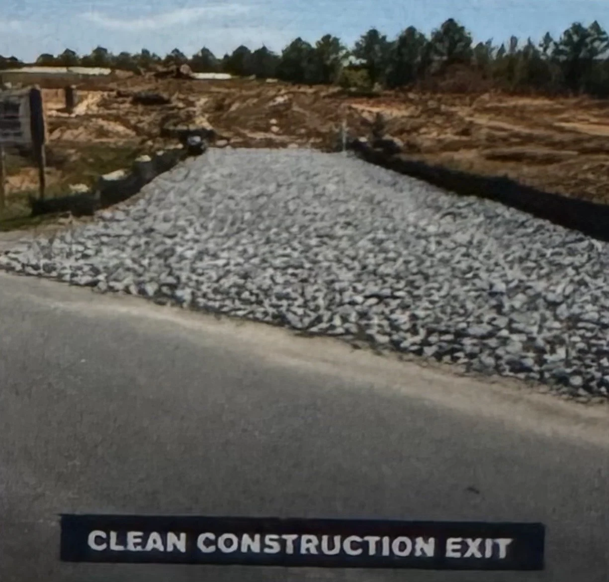 A gravel construction exit area with a sign that reads 'CLEAN CONSTRUCTION EXIT' and a landscape in the background with dirt mounds and trees.