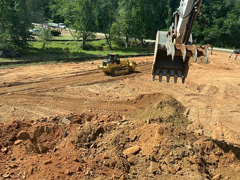 Construction site with an excavator's bucket digging into dirt, a yellow bulldozer in the background, and a line of trees and parked vehicles beyond.