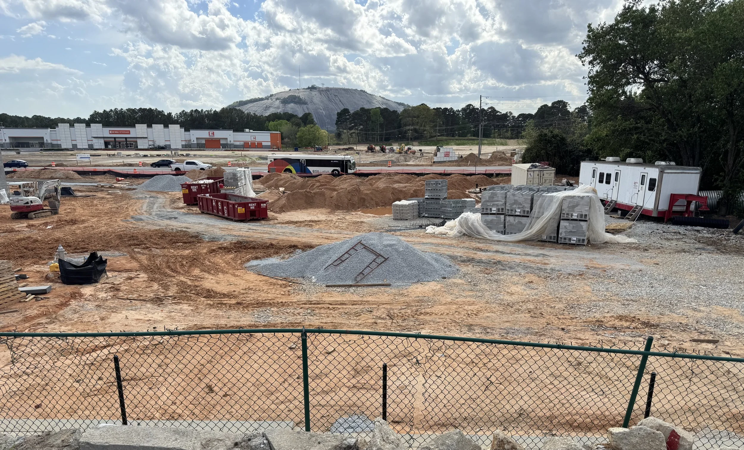 Construction site with piles of dirt, construction equipment, and building materials. Backdrop features a large hill or mountain with some structures on top, partly cloudy sky, and nearby trees.