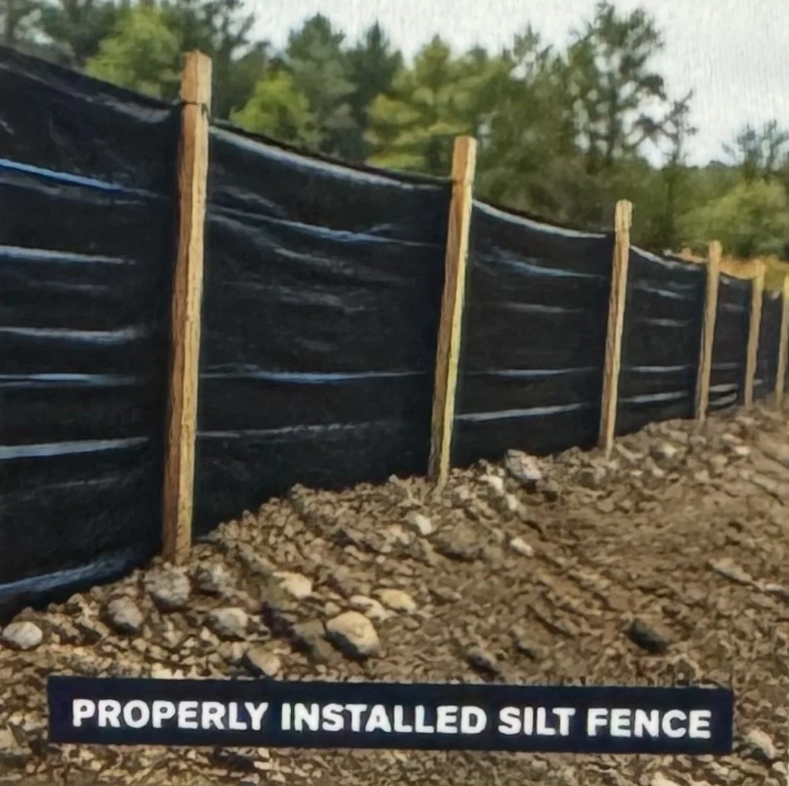 A black silt fence installed along a rocky ground with wooden posts, with trees and blue sky in the background.