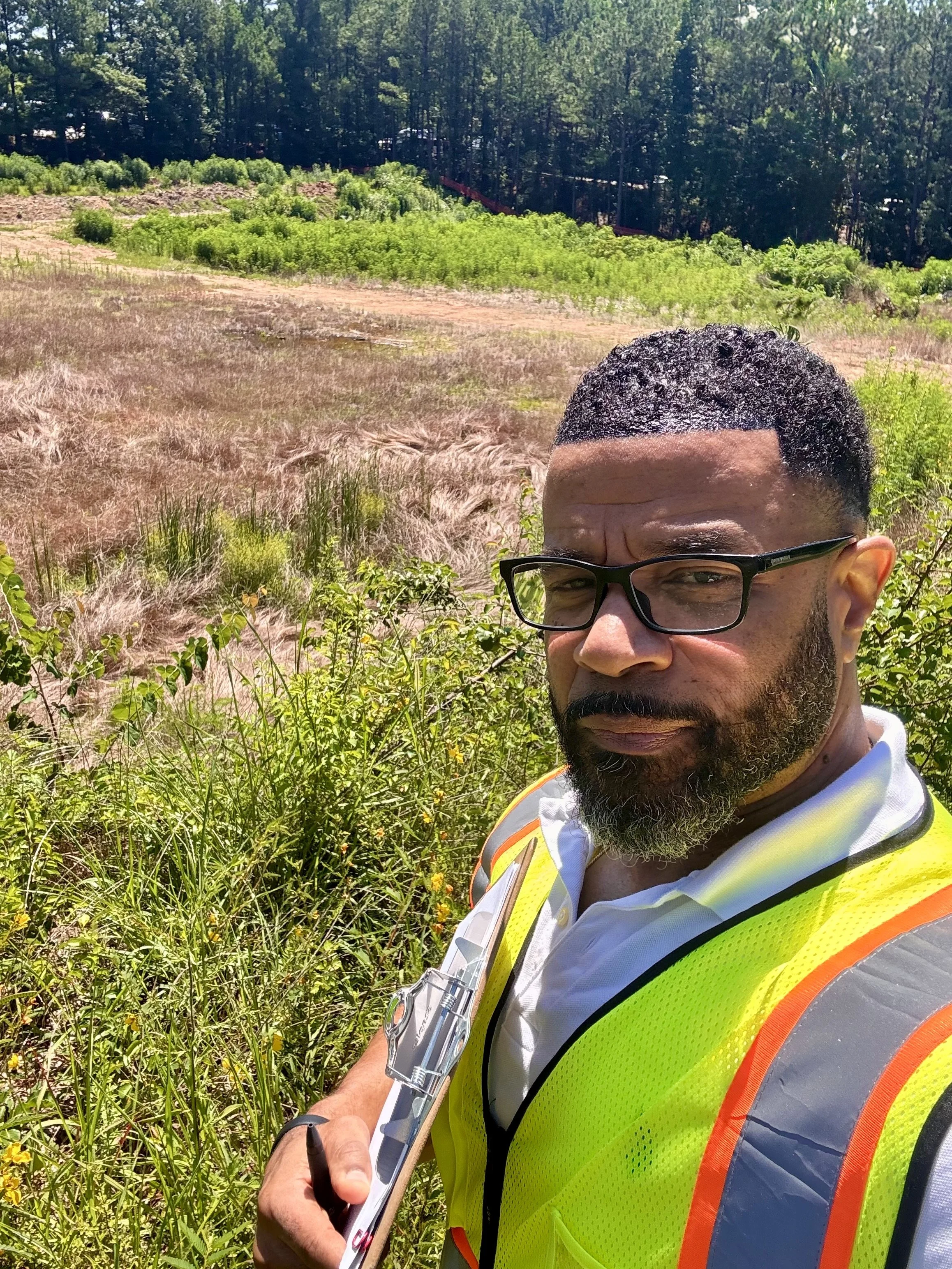 A man wearing glasses and a yellow safety vest taking notes outdoors near tall grass and shrubs. In the background, there are trees and a pathway.