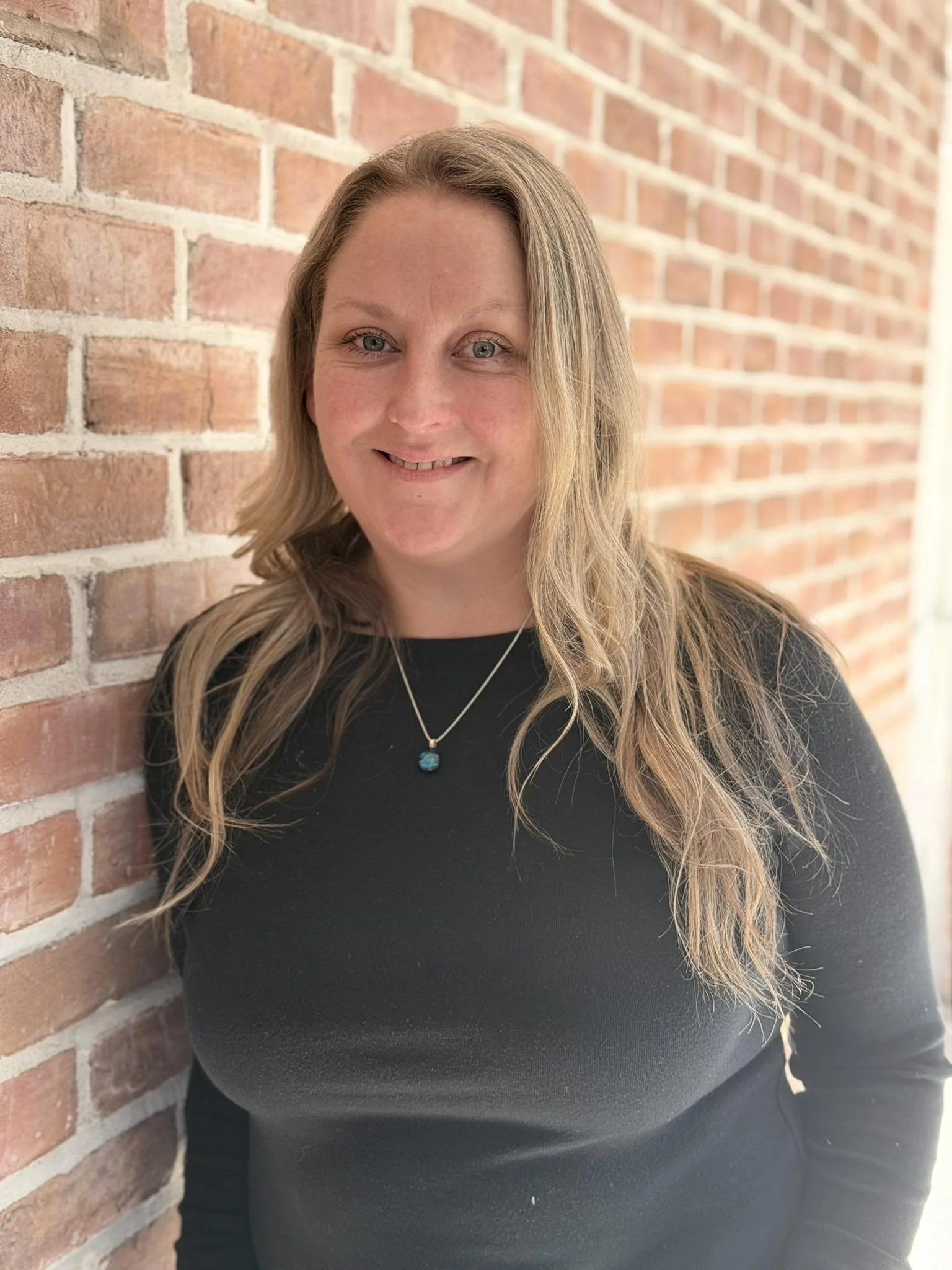 A woman with long, wavy blonde hair and blue eyes leaning against a brick wall, smiling at the camera, wearing a black top and a necklace with a blue pendant.