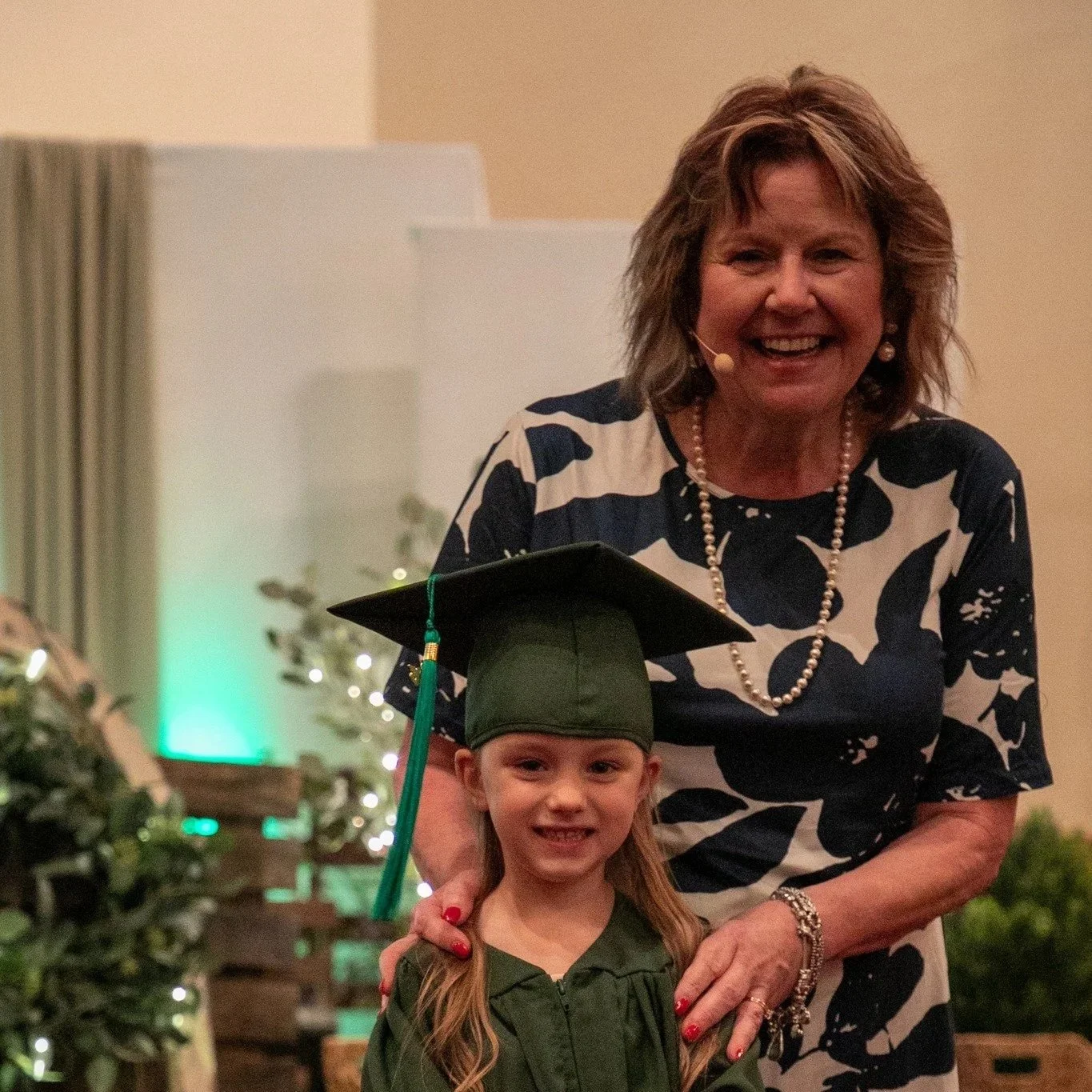 A young girl in a green graduation cap and gown standing next to an older woman, both smiling, during a graduation ceremony.