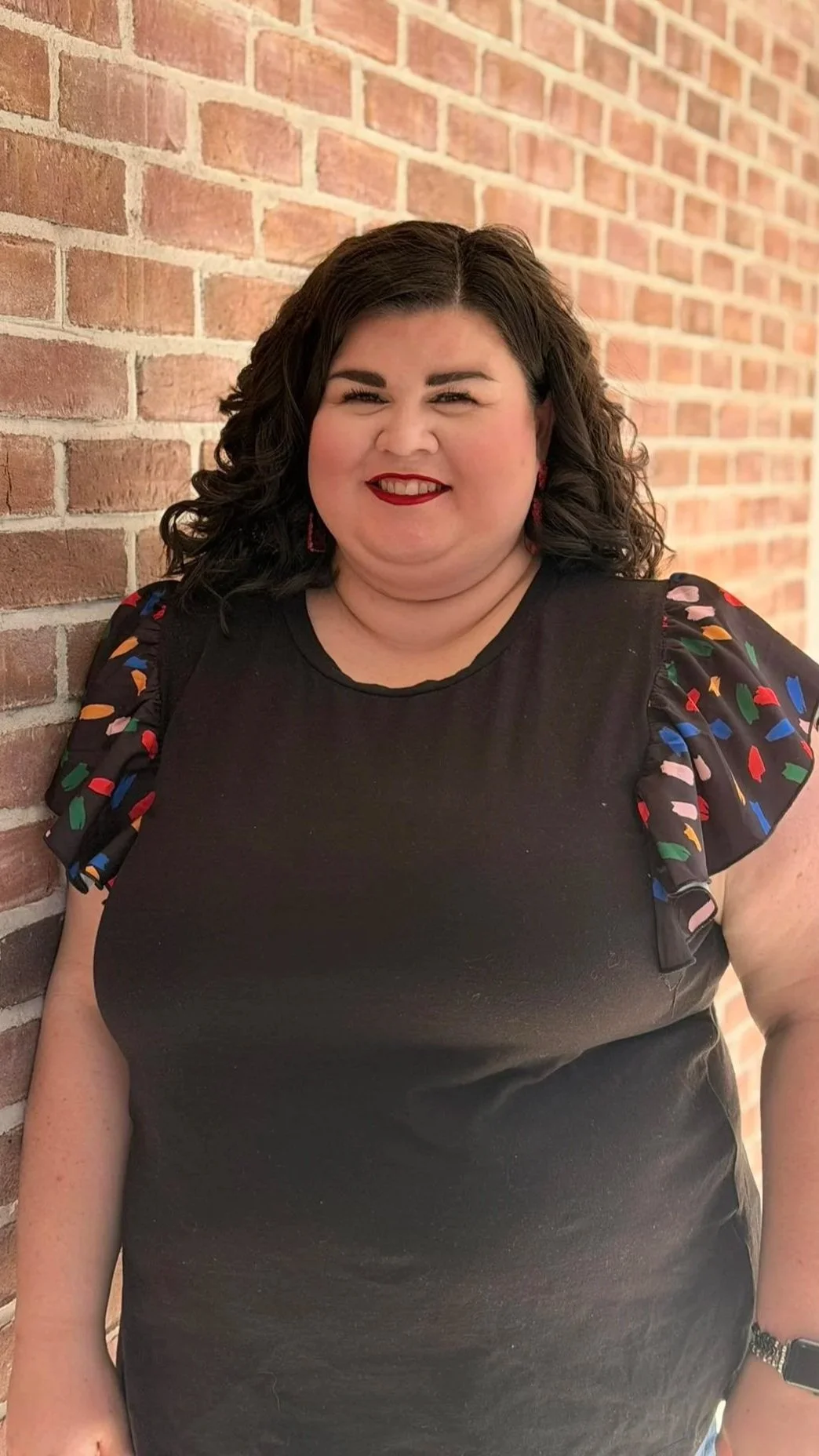 A woman with dark curly hair, red lipstick, and a black dress with colorful puffed sleeves, smiling while standing against a brick wall.